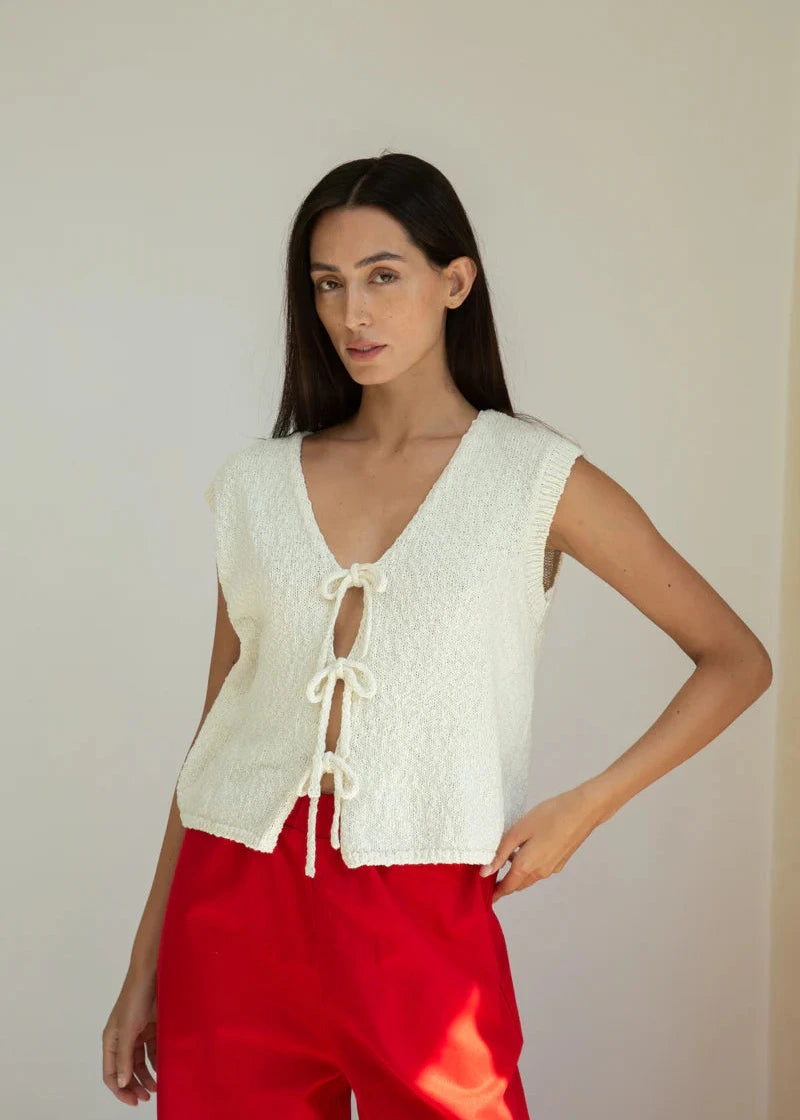 Woman wearing a white sleeveless top with lace-up front and red pants against a plain background