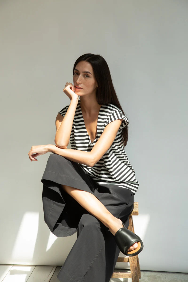 Woman in a black and white striped top and gray pants sitting on a wooden stool against a plain wall.