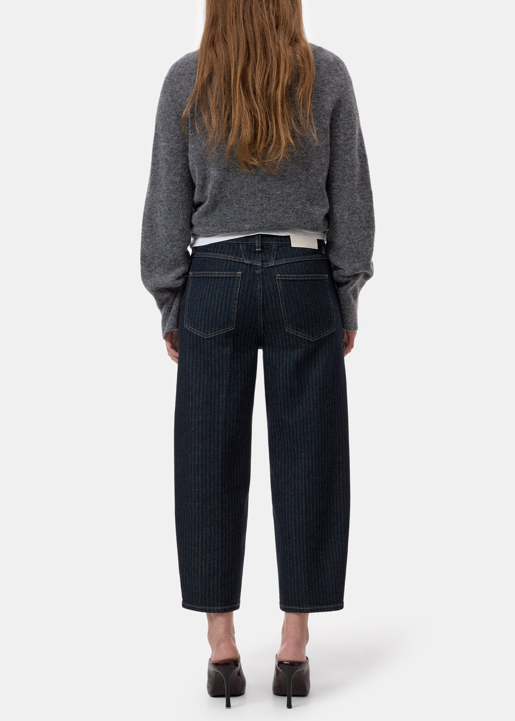 A model shown from the back wearing dark navy pinstriped cropped jeans, a grey knit sweater, and black heels against a light gray studio background.