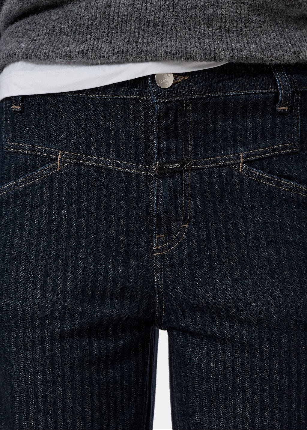 Close-up of dark indigo denim jeans with a subtle vertical stripe texture, showing the front pocket details, belt loops, and a white t-shirt tucked in, paired with a gray knit sweater.