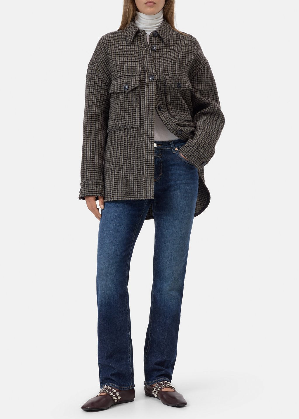 A model wearing a brown and black checkered button-up shirt jacket over a white turtleneck, paired with blue jeans and brown studded loafers, on a light gray background.