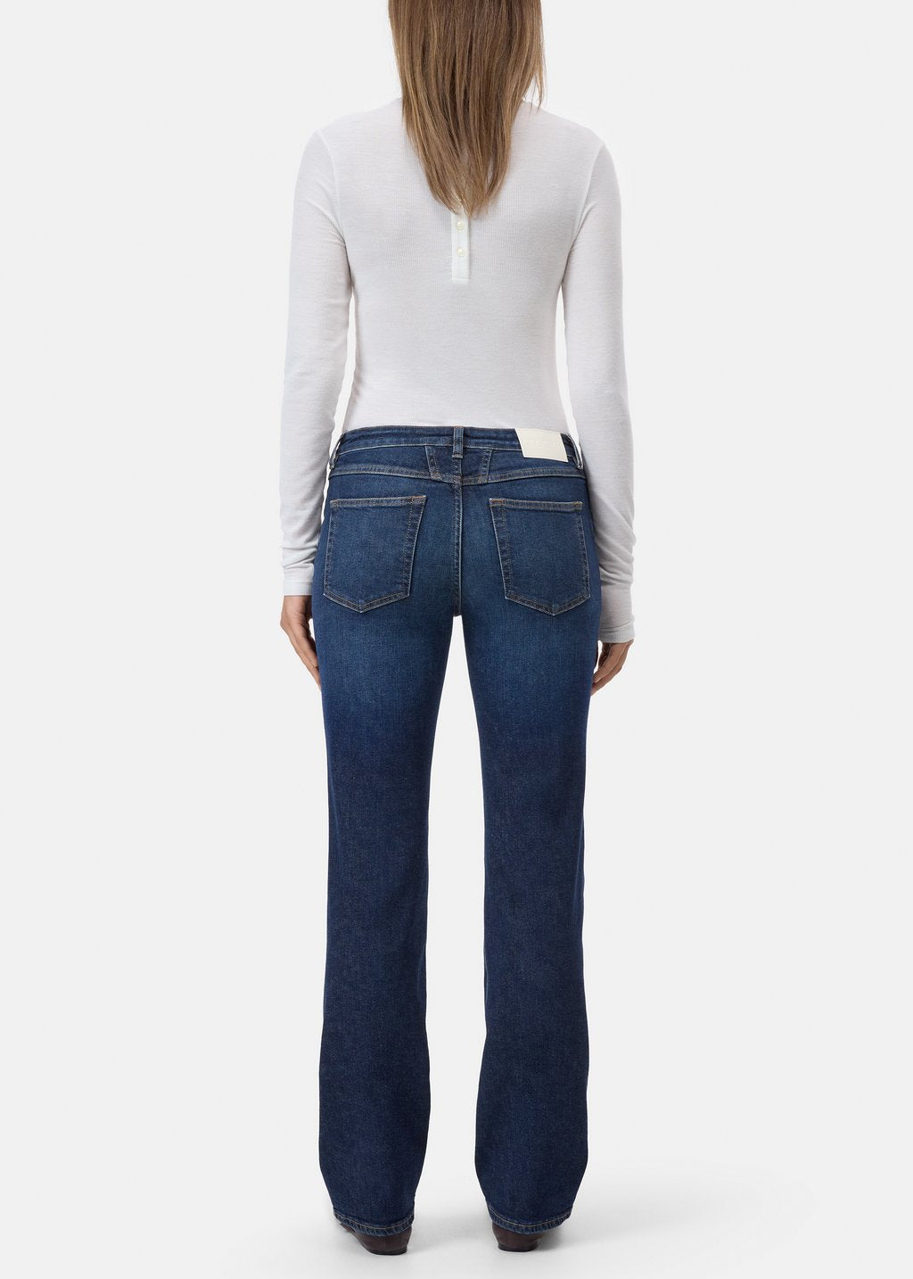A woman viewed from the back wearing a white long-sleeve top and dark blue straight-leg jeans against a light gray studio background.