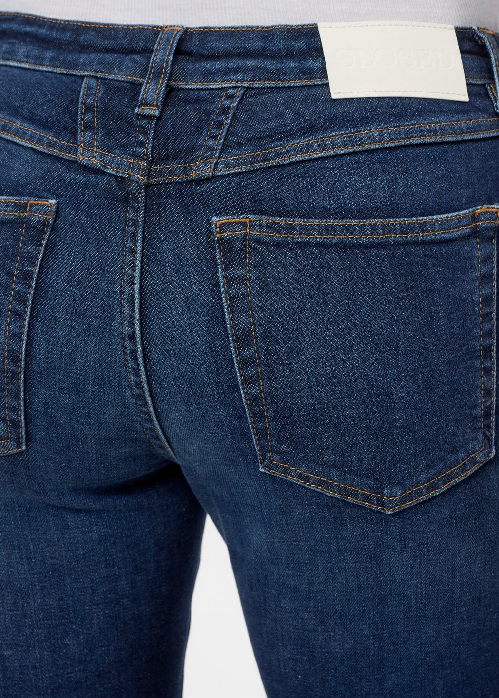 Close-up back view of dark blue denim jeans on a model, showing the waistband, belt loops, back pockets, and a white brand label.