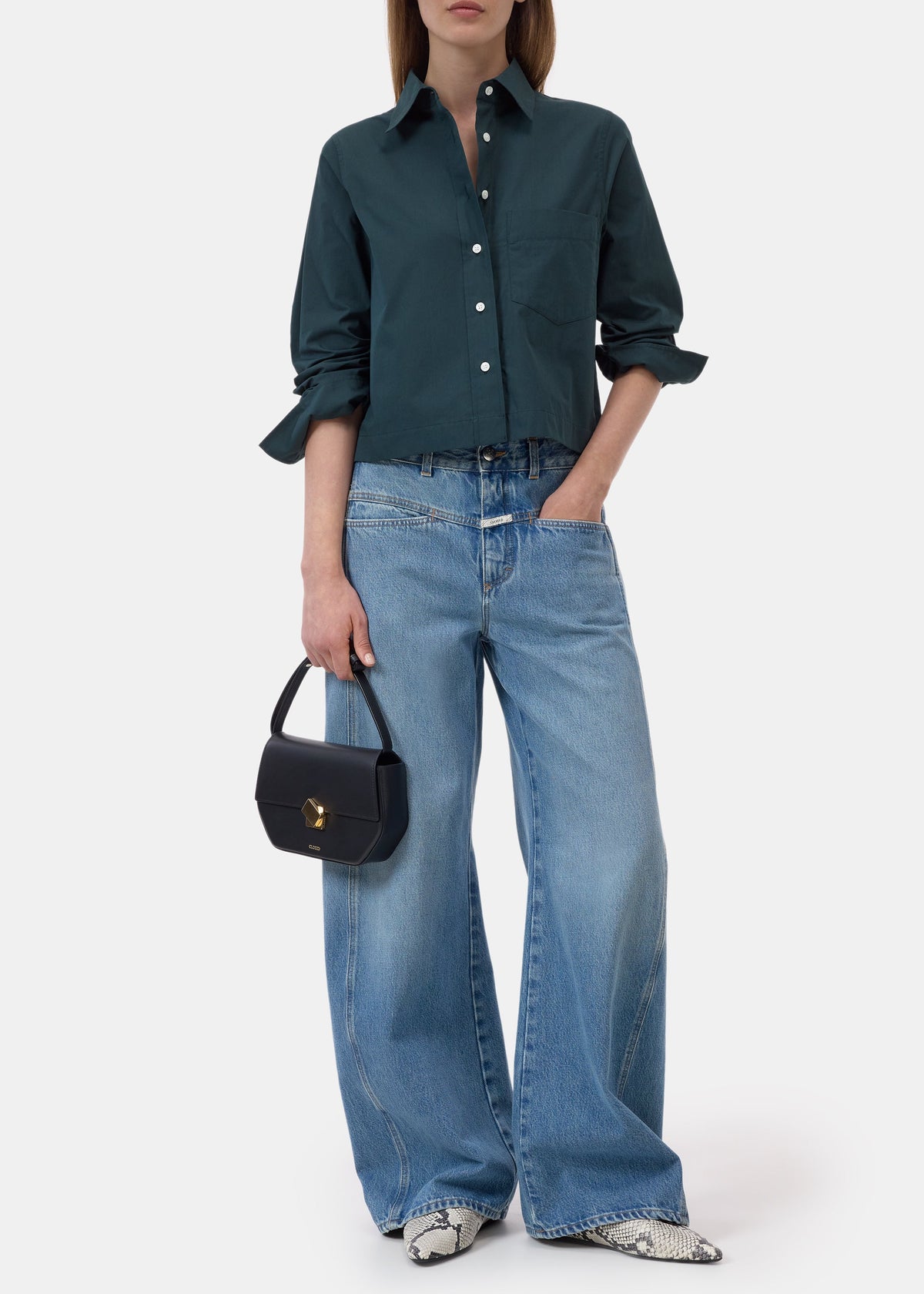 A model wearing a dark teal button-up shirt, wide-leg light blue jeans, and snakeskin-patterned shoes, holding a small black handbag against a plain white studio background.