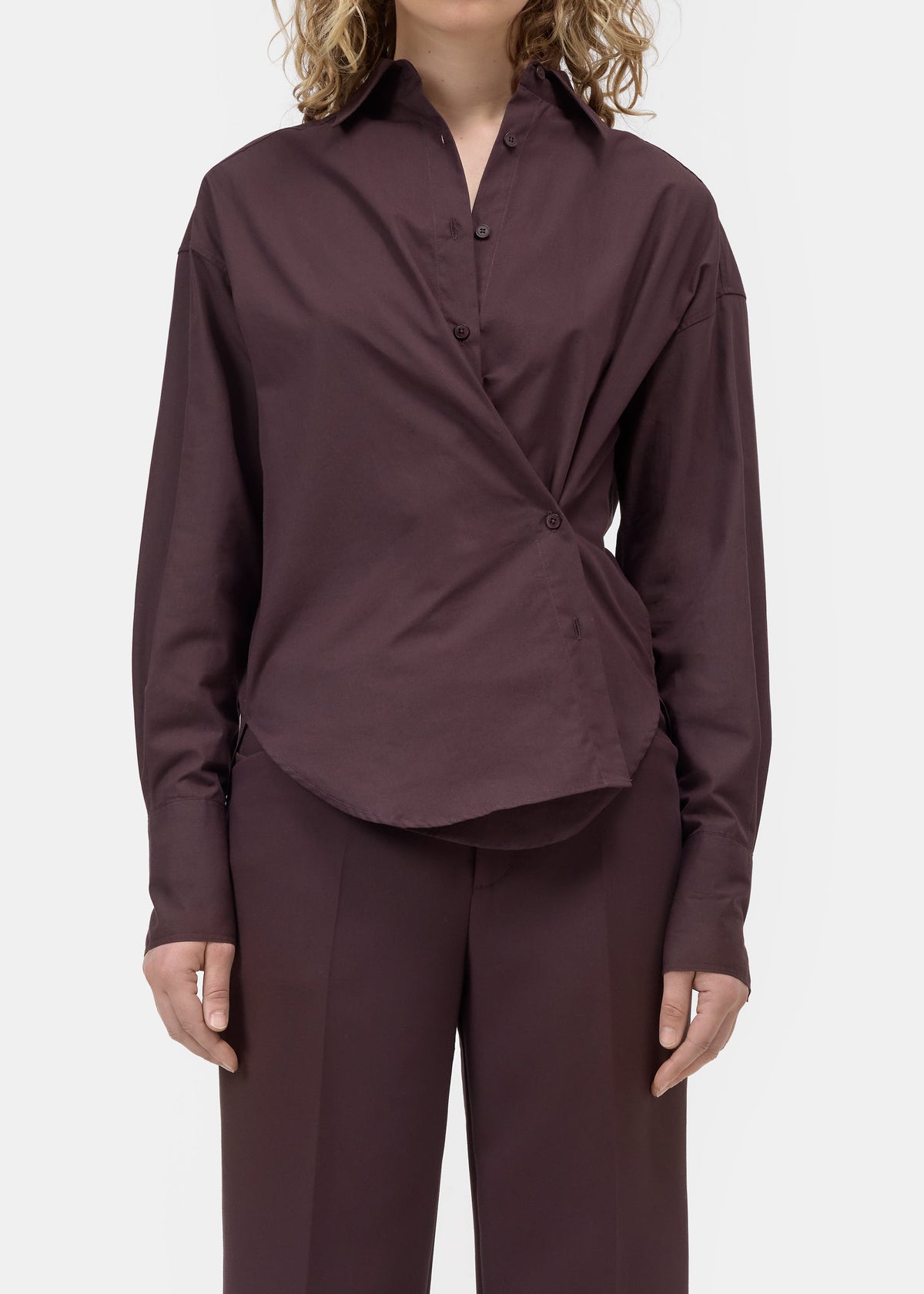A model wearing a dark plum long-sleeve button-up shirt with a unique twisted front detail and matching dark plum trousers, shot against a light gray studio background.