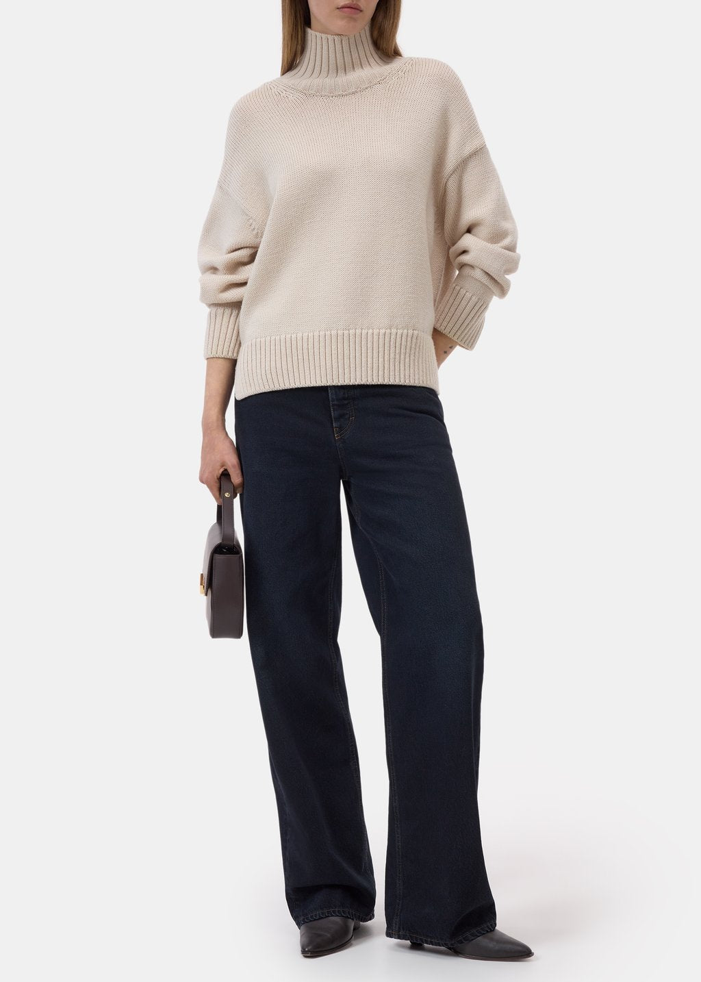 A model wearing a cream-colored ribbed turtleneck sweater and dark navy wide-leg jeans, holding a small dark handbag against a plain light gray studio background.