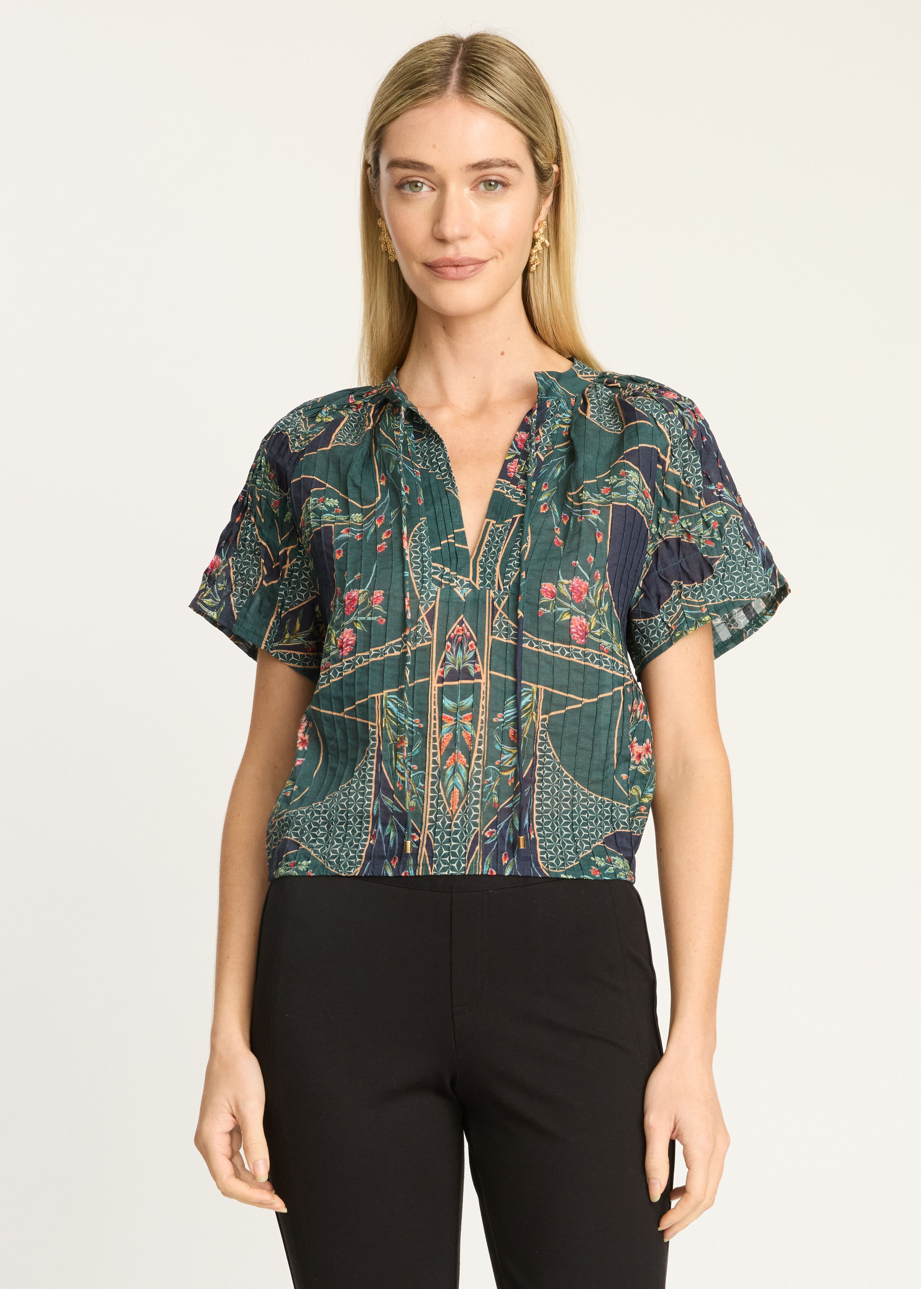 A woman modeling a short-sleeve button-front blouse with a dark green and navy blue abstract pattern, paired with black pants, against a plain light background.