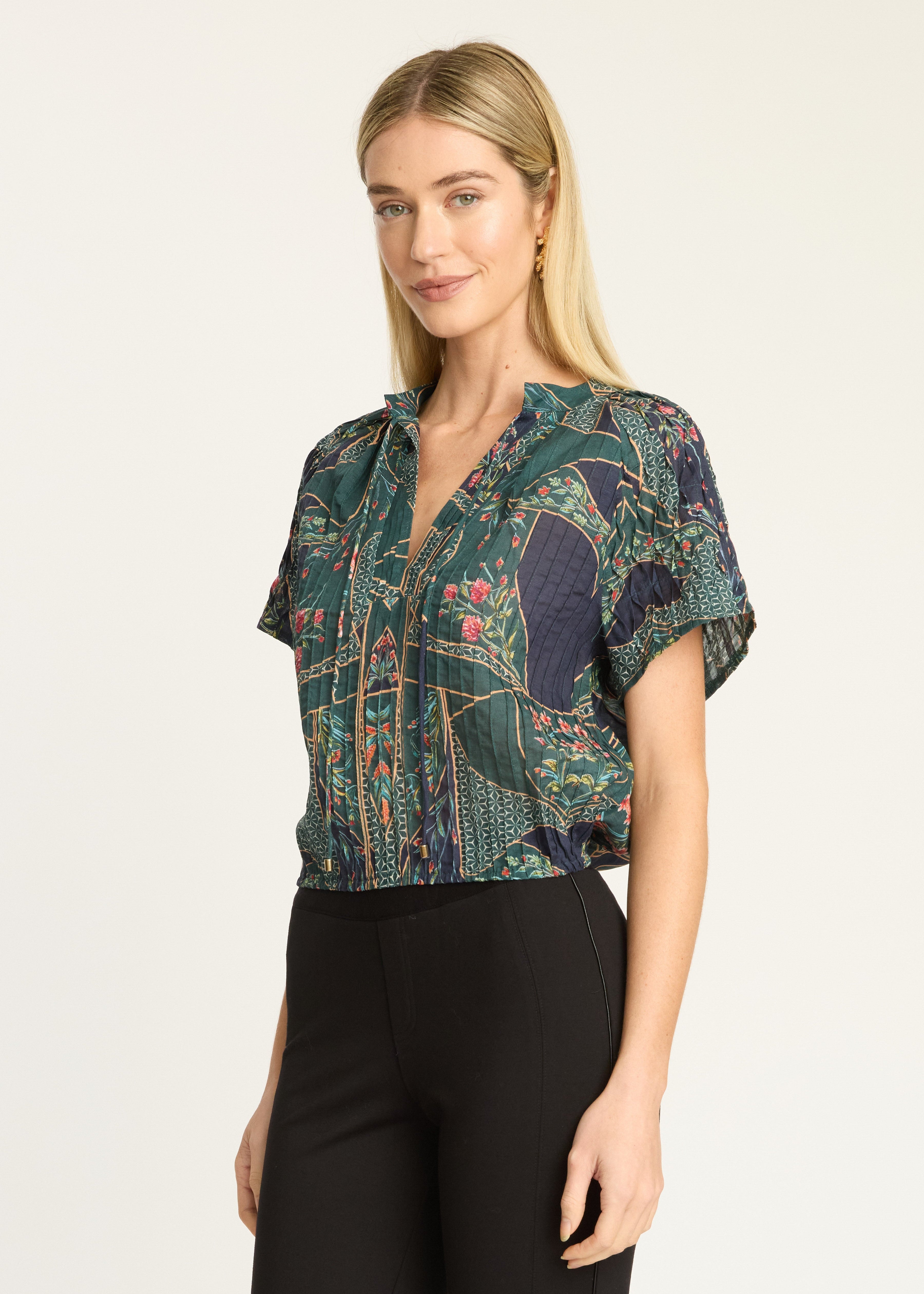 A woman modeling a short-sleeve button-up blouse with a dark green and navy blue botanical pattern, paired with black pants, against a plain light background.