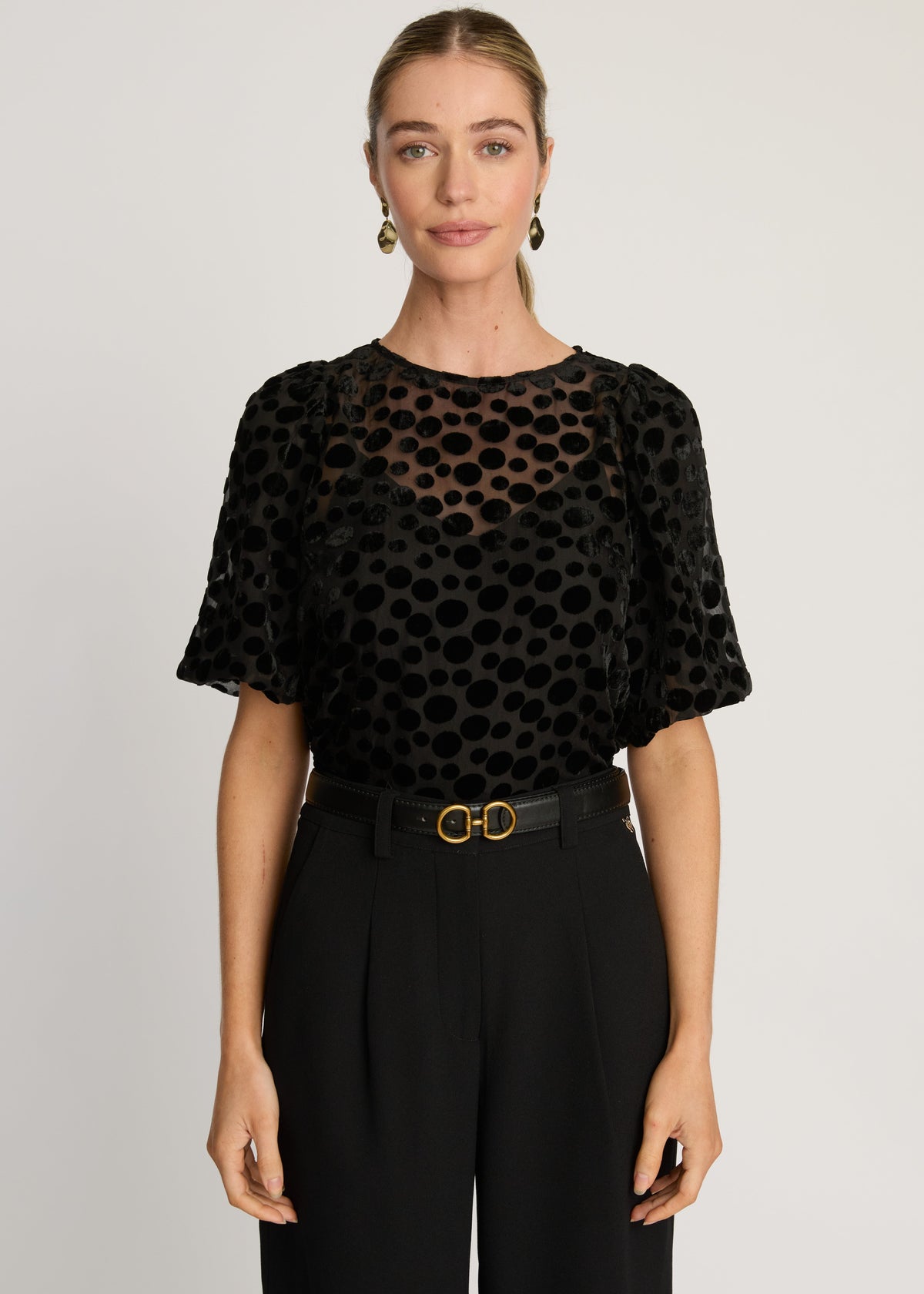 Woman wearing a black patterned top and black pants on a plain background