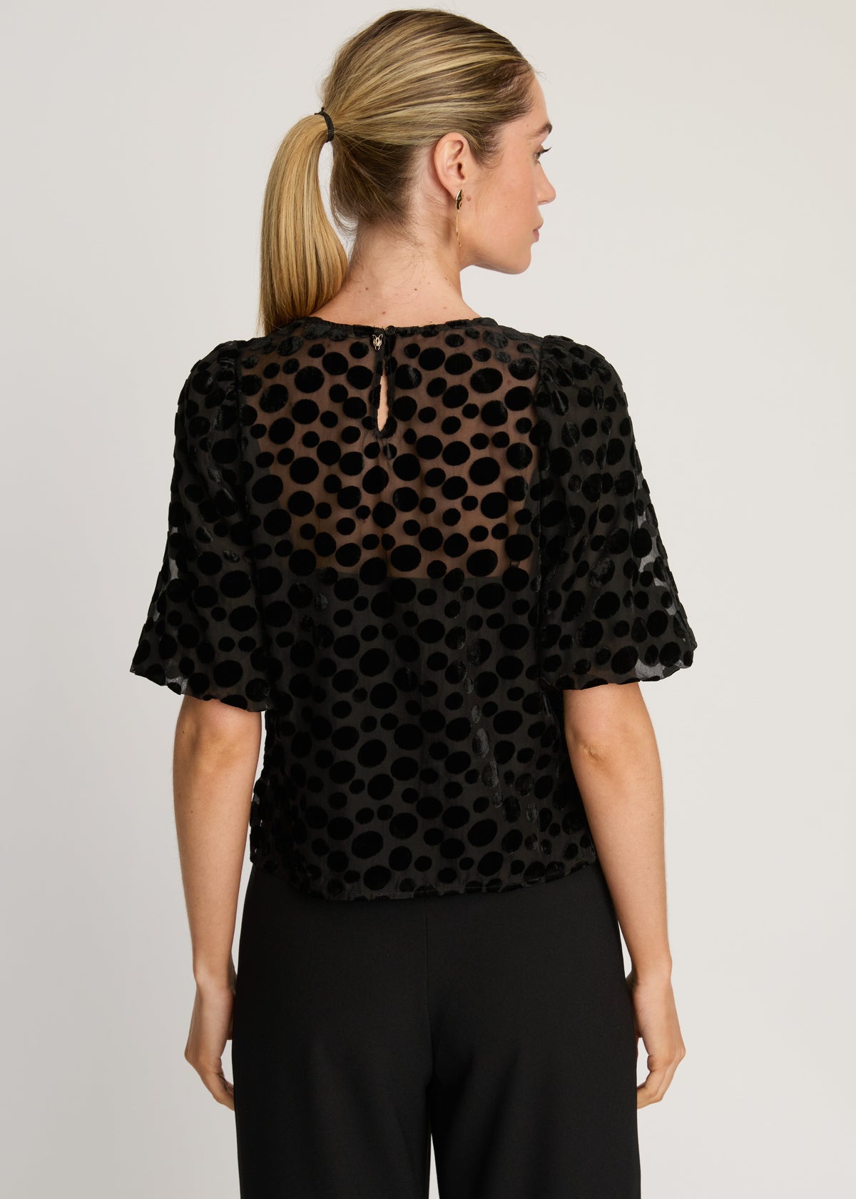 Woman wearing a black polka dot blouse with sheer sleeves on a plain background