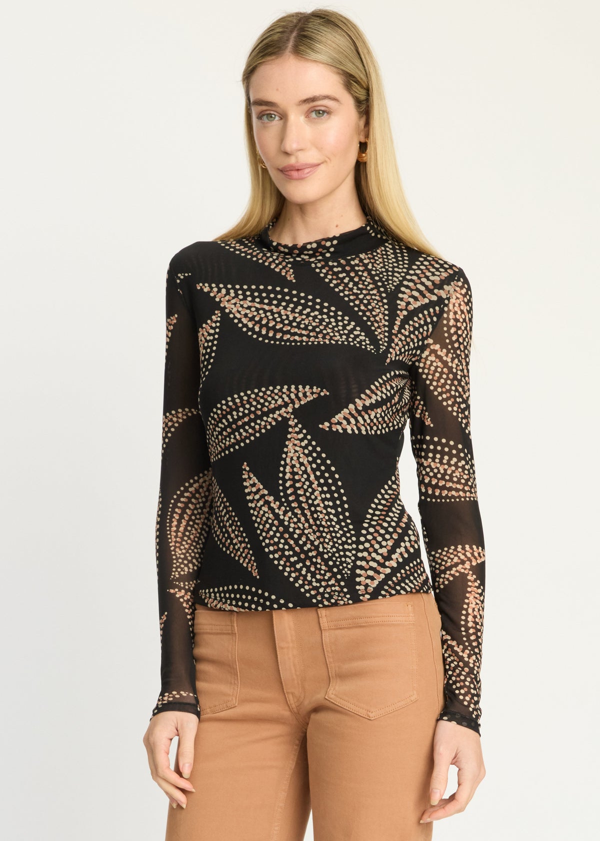 A woman modeling a black long-sleeve top with a beige leaf pattern, paired with tan pants, against a plain light gray background.