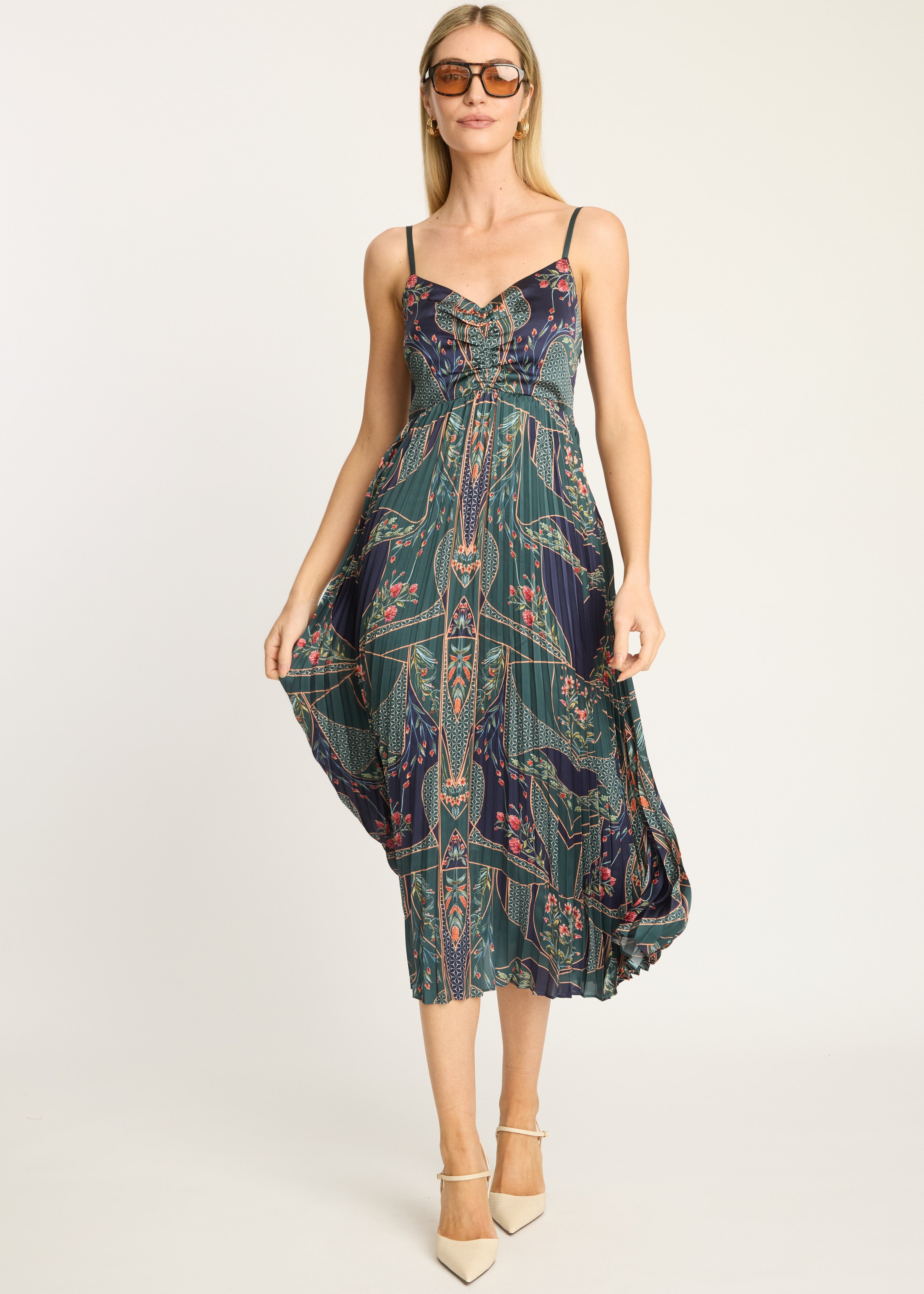 A woman modeling a sleeveless midi dress with thin straps and a dark teal, green, and navy paisley print, styled with beige heels and sunglasses against a plain light background.