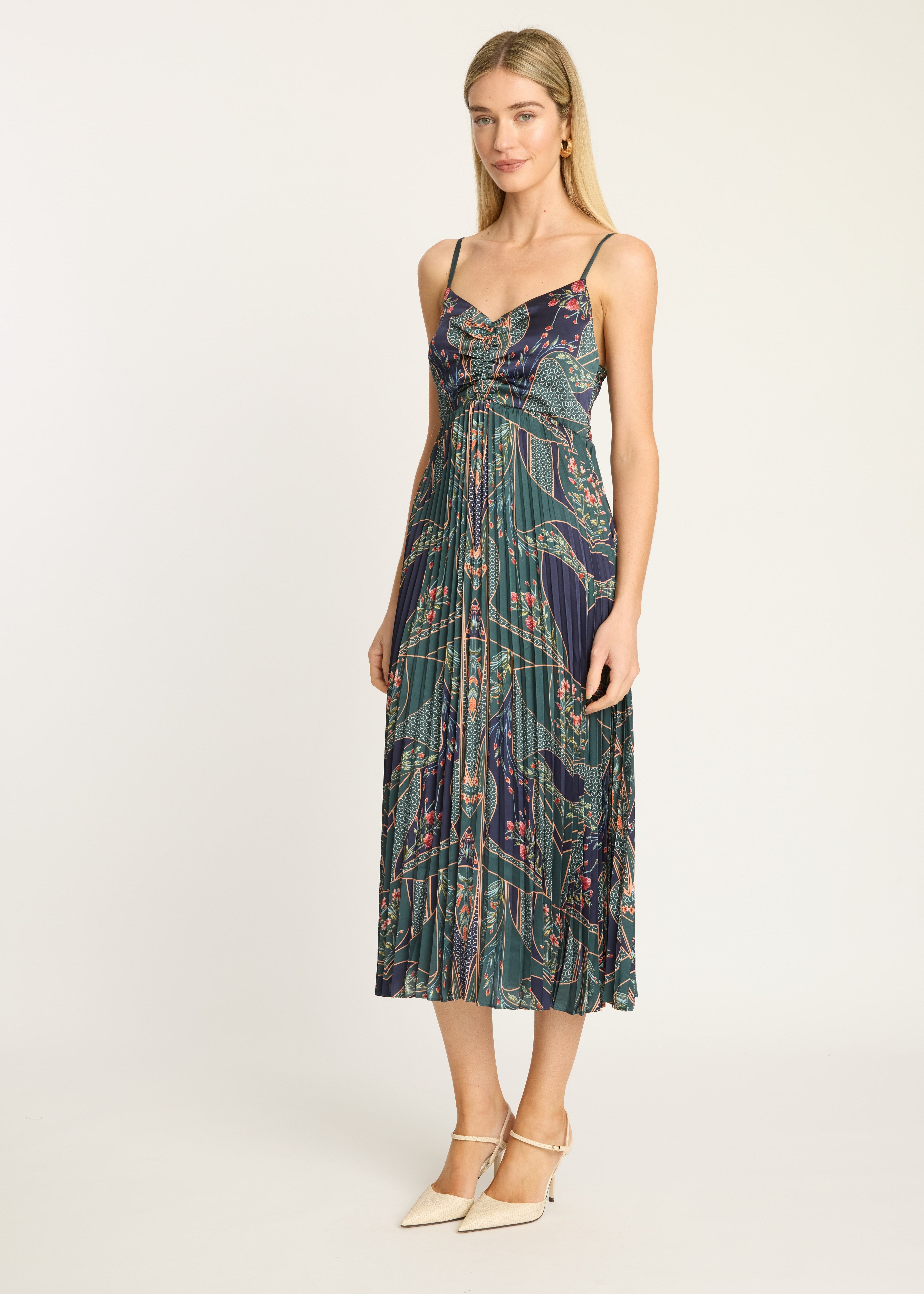 A woman modeling a sleeveless midi dress with thin straps and a gathered sweetheart neckline, featuring an abstract botanical print in dark teal, green, purple, and pink. She is wearing cream-colored pointed-toe heels against a plain light gray studio background.