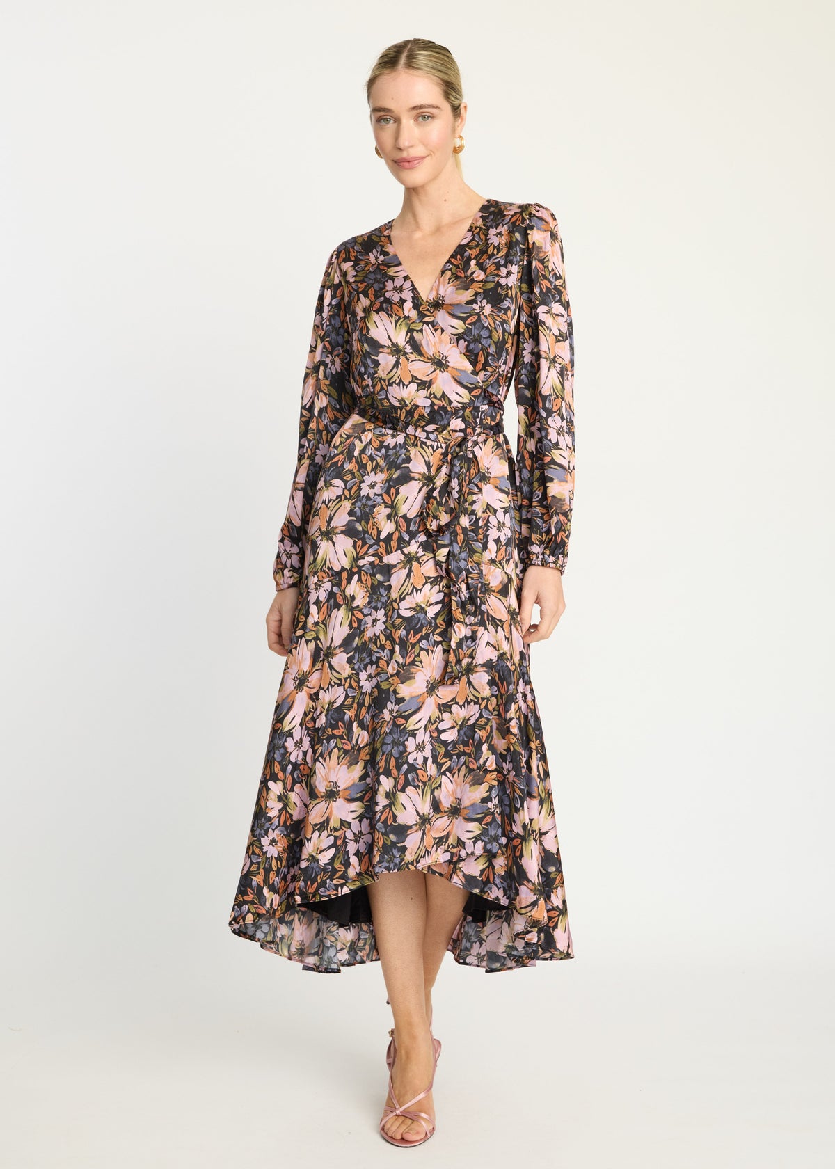 A woman modeling a long-sleeve floral midi dress with a wrap-style V-neck, tie waist, and high-low hem, photographed against a plain light gray studio background.