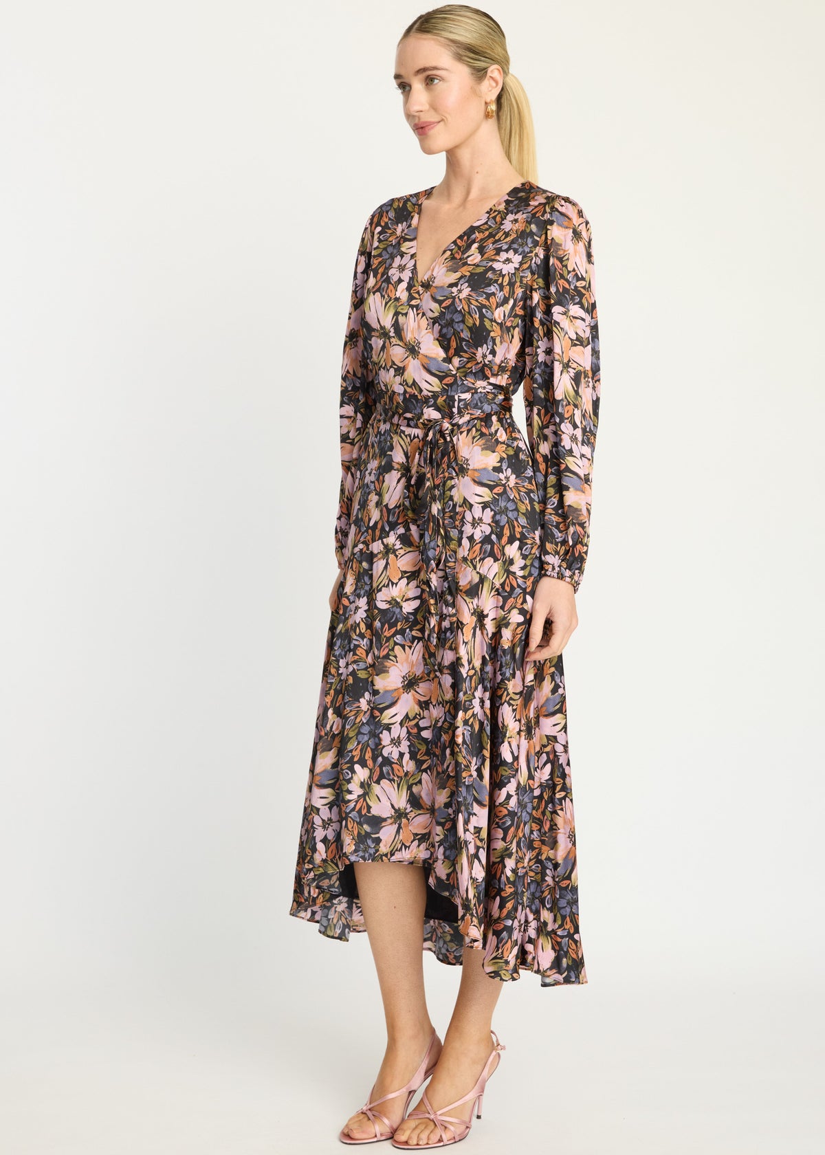A woman modeling a long-sleeve midi wrap dress with a dense floral print in pink, blue, and beige tones, paired with light pink strappy heels, against a plain white studio background.