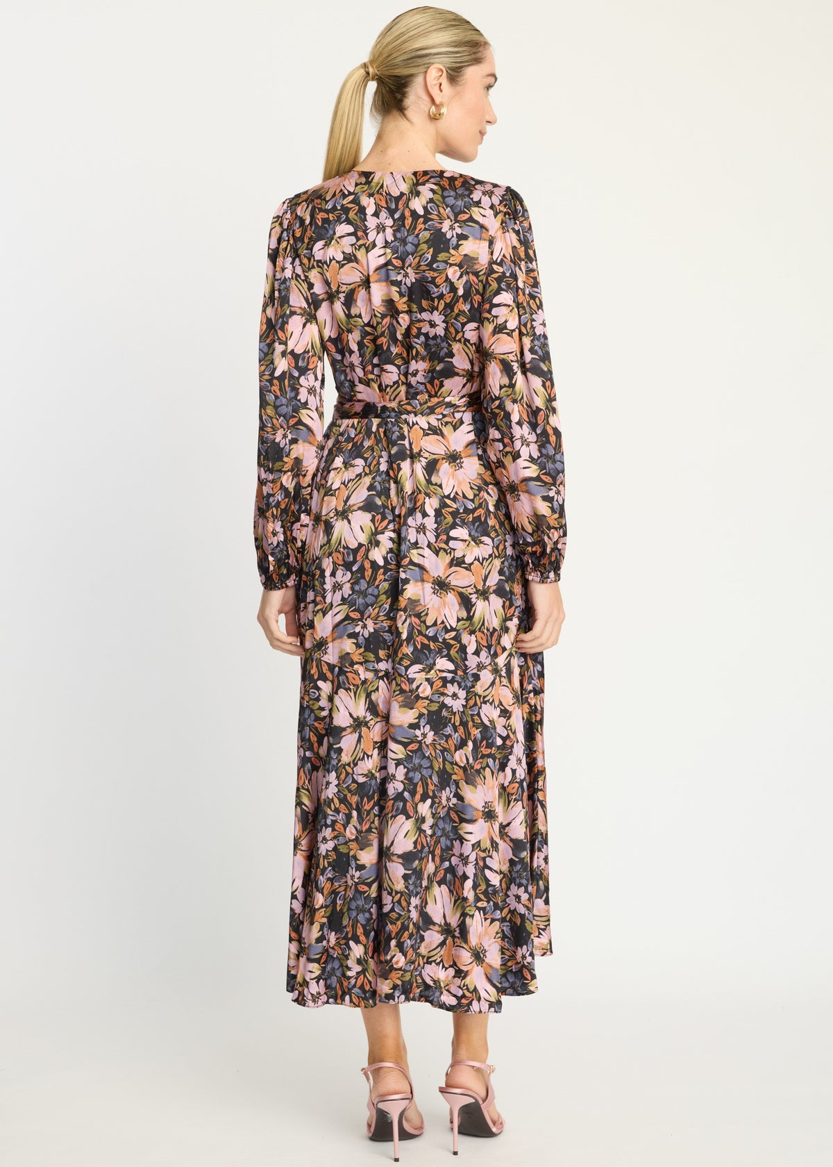 A woman viewed from the back wearing a long-sleeve midi dress with a dense floral print in pink, beige, and dark navy/black tones, paired with pink high-heeled sandals against a light gray studio background.