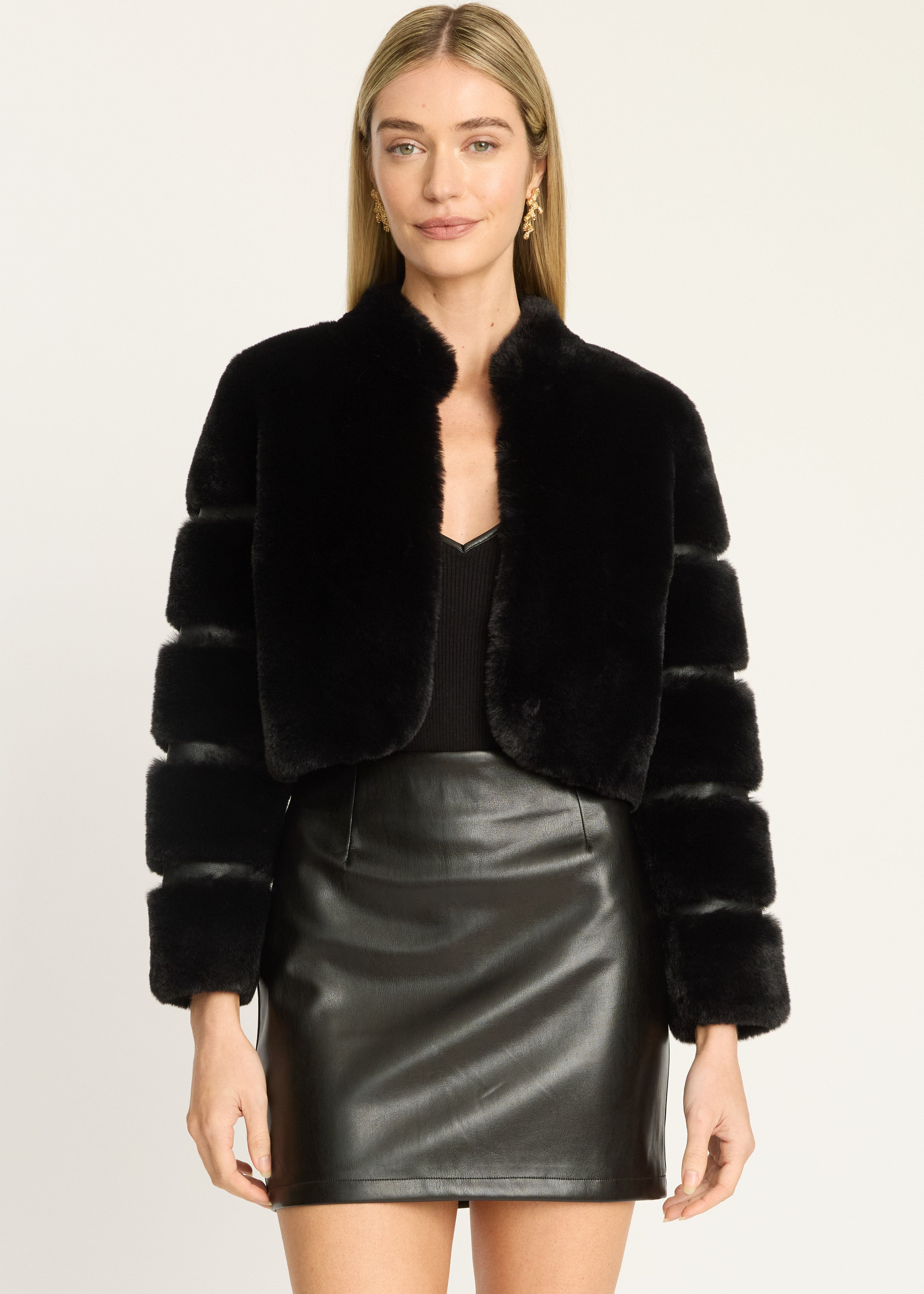 A woman modeling a black cropped faux-fur jacket with ribbed sleeves, paired with a black top and a black faux-leather mini skirt, against a plain light background.