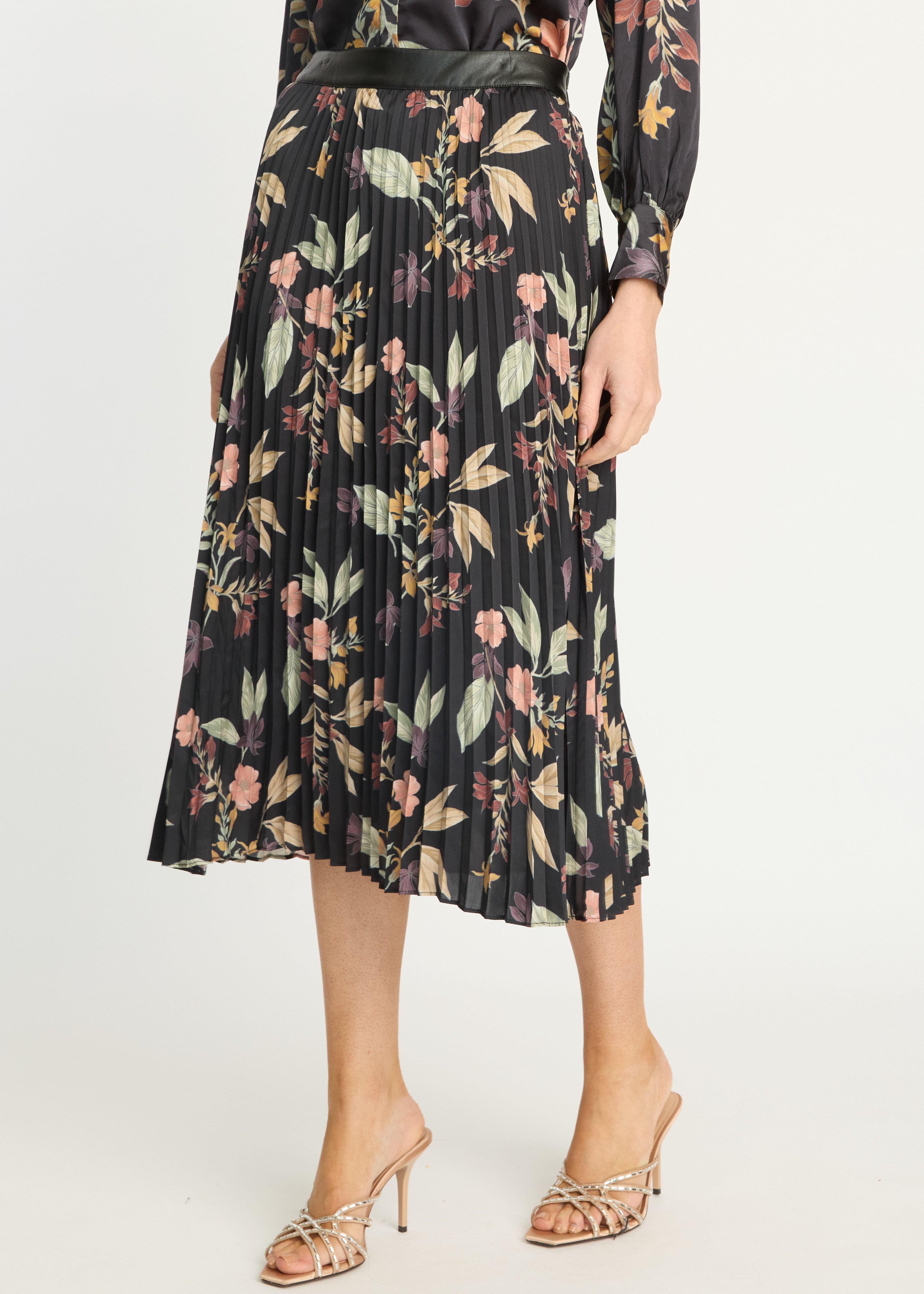A model wearing a black pleated midi skirt with a multicolor floral print, paired with a black belt and metallic heeled sandals, against a plain light background.