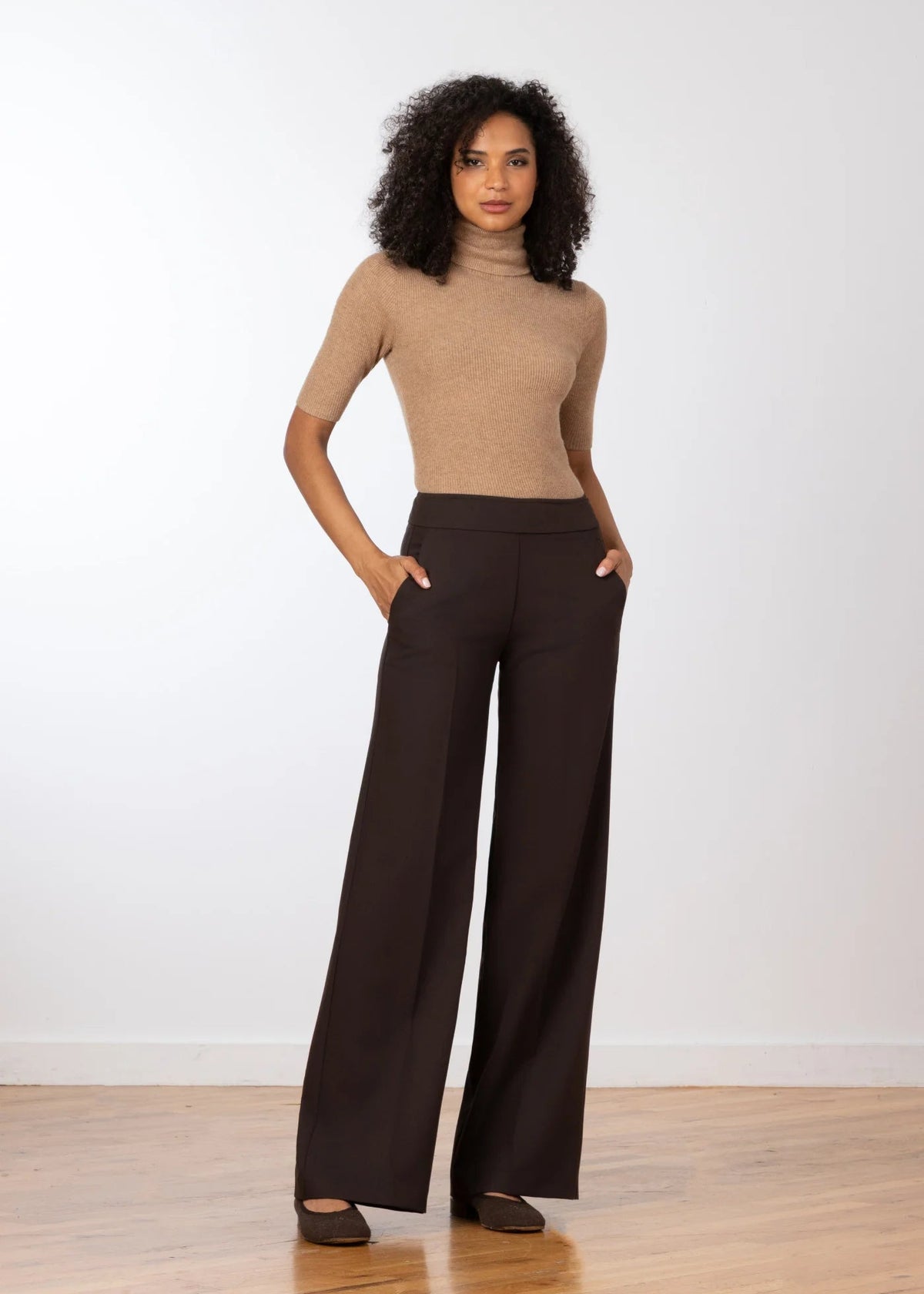 A woman stands in a studio wearing a fitted beige short-sleeve turtleneck top and dark brown wide-leg trousers, with her hands in the trouser pockets. She is posed on a light wood floor against a plain white wall.