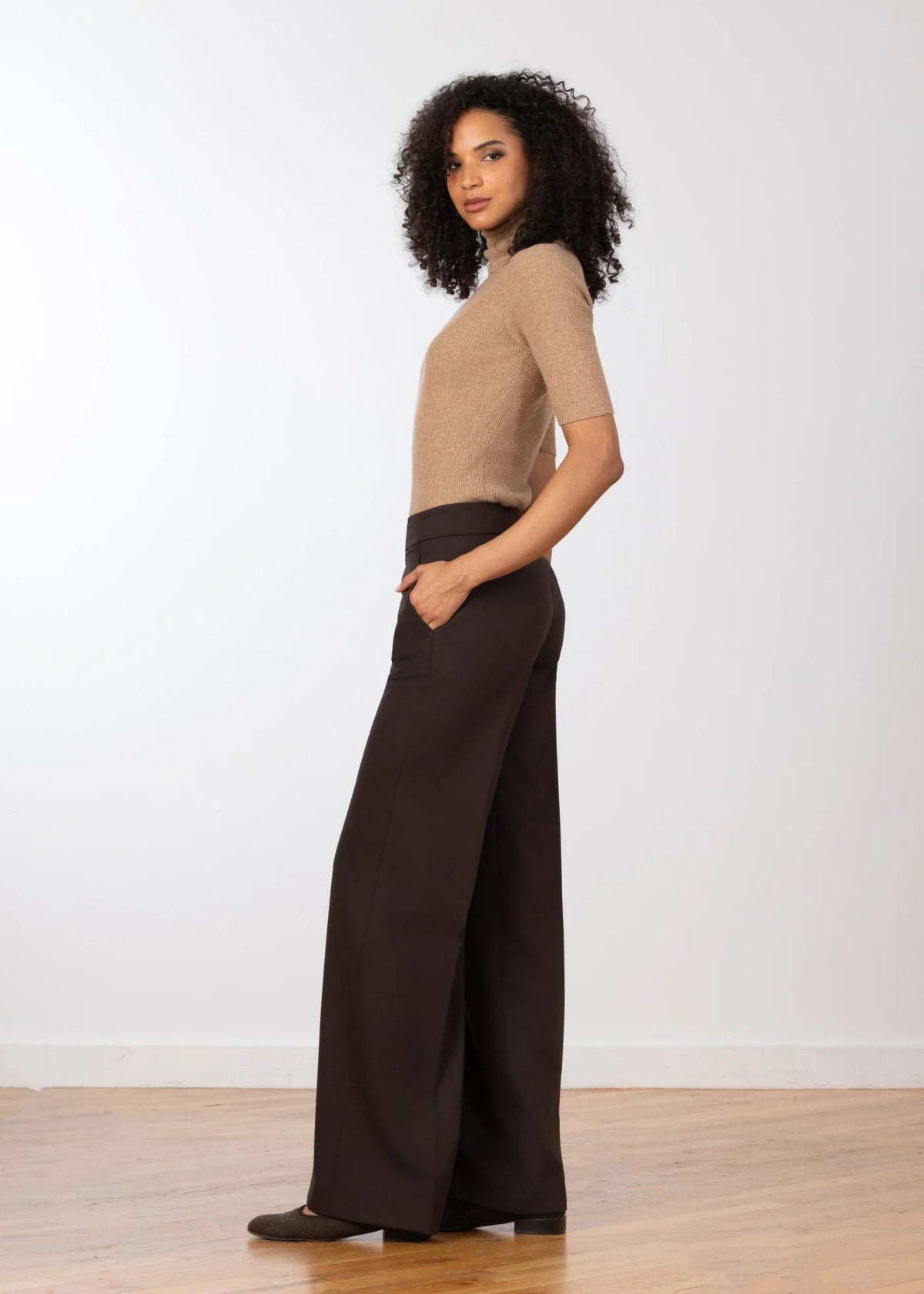 A woman stands in profile wearing a beige short-sleeve knit top and dark brown wide-leg trousers, standing on a light wood floor against a plain white wall.