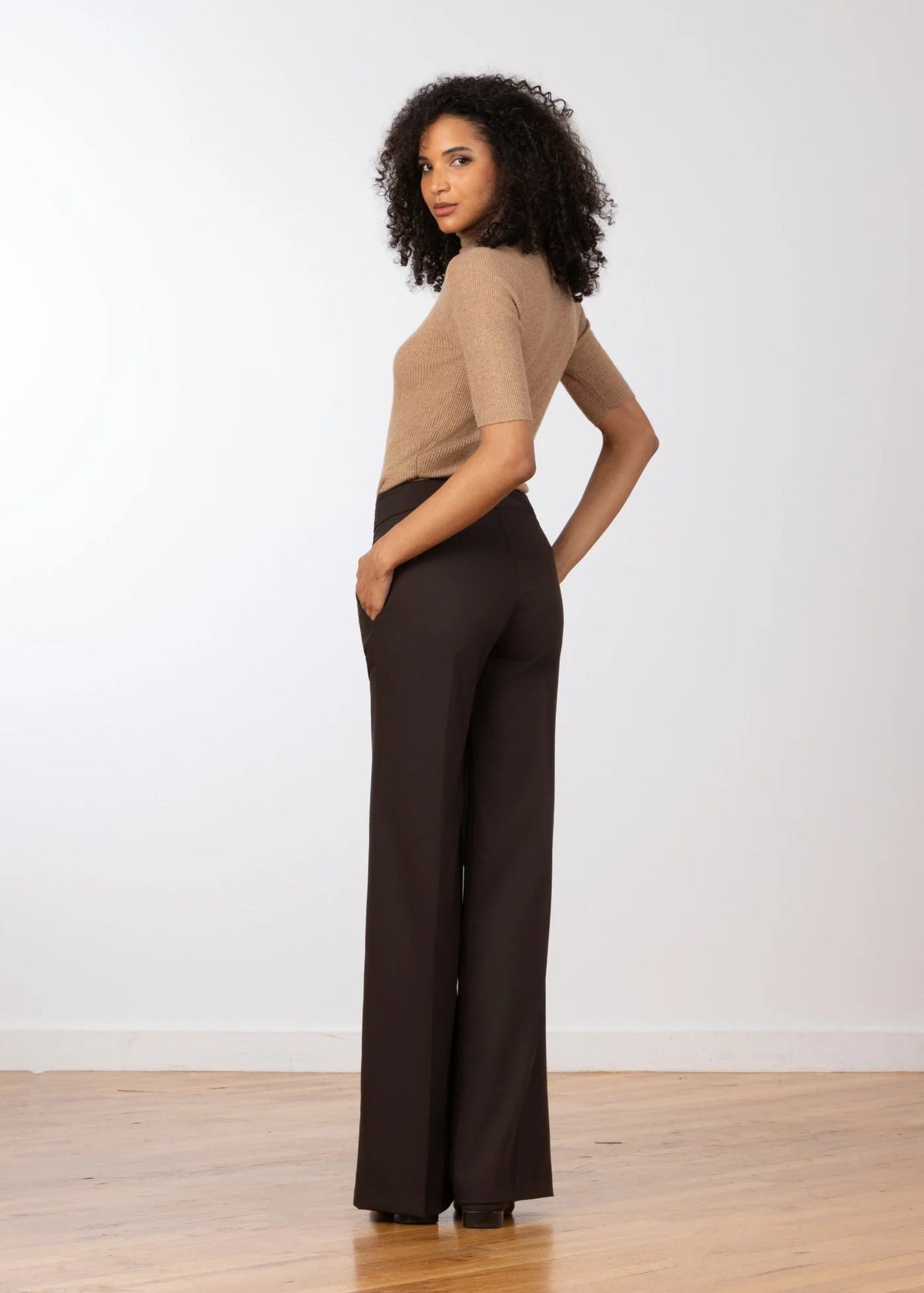 A woman stands in profile wearing a beige short-sleeve knit top and dark brown wide-leg trousers against a plain white wall and light wood floor.