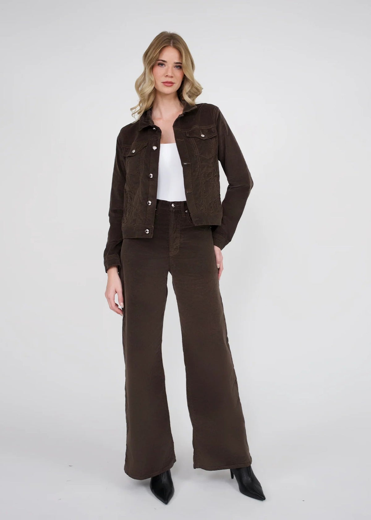 A woman modeling a dark brown corduroy jacket and matching wide-leg pants over a white top, standing against a light gray studio background.