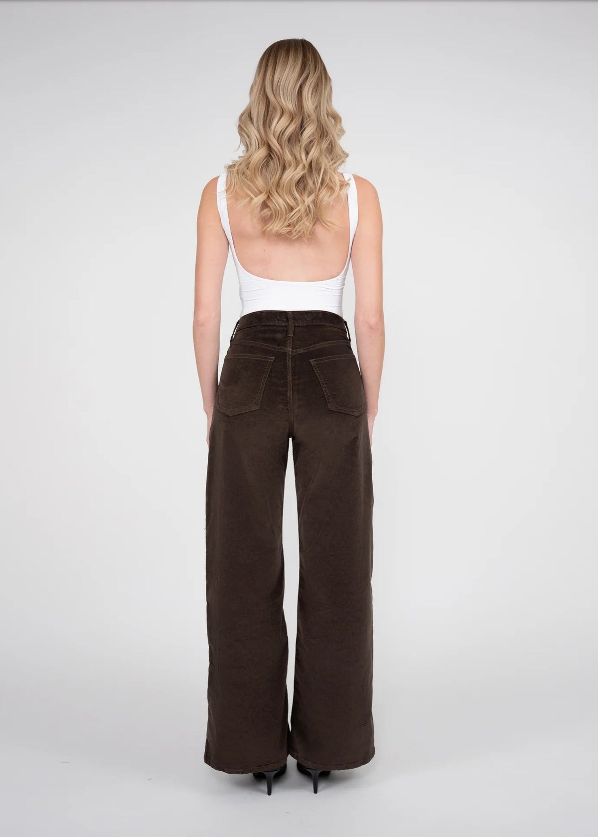 A woman is shown from behind wearing high-waisted, wide-leg brown corduroy pants and a white tank top against a light gray studio background.