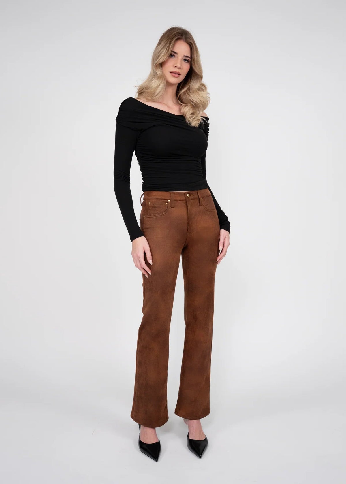 A woman modeling a black off-the-shoulder long-sleeve top and brown wide-leg pants with black pointed-toe heels, standing against a light gray studio background.