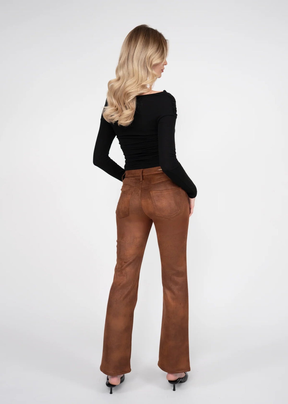 A woman is shown from behind wearing brown flared pants and a black long-sleeve top, standing against a plain light gray studio background.