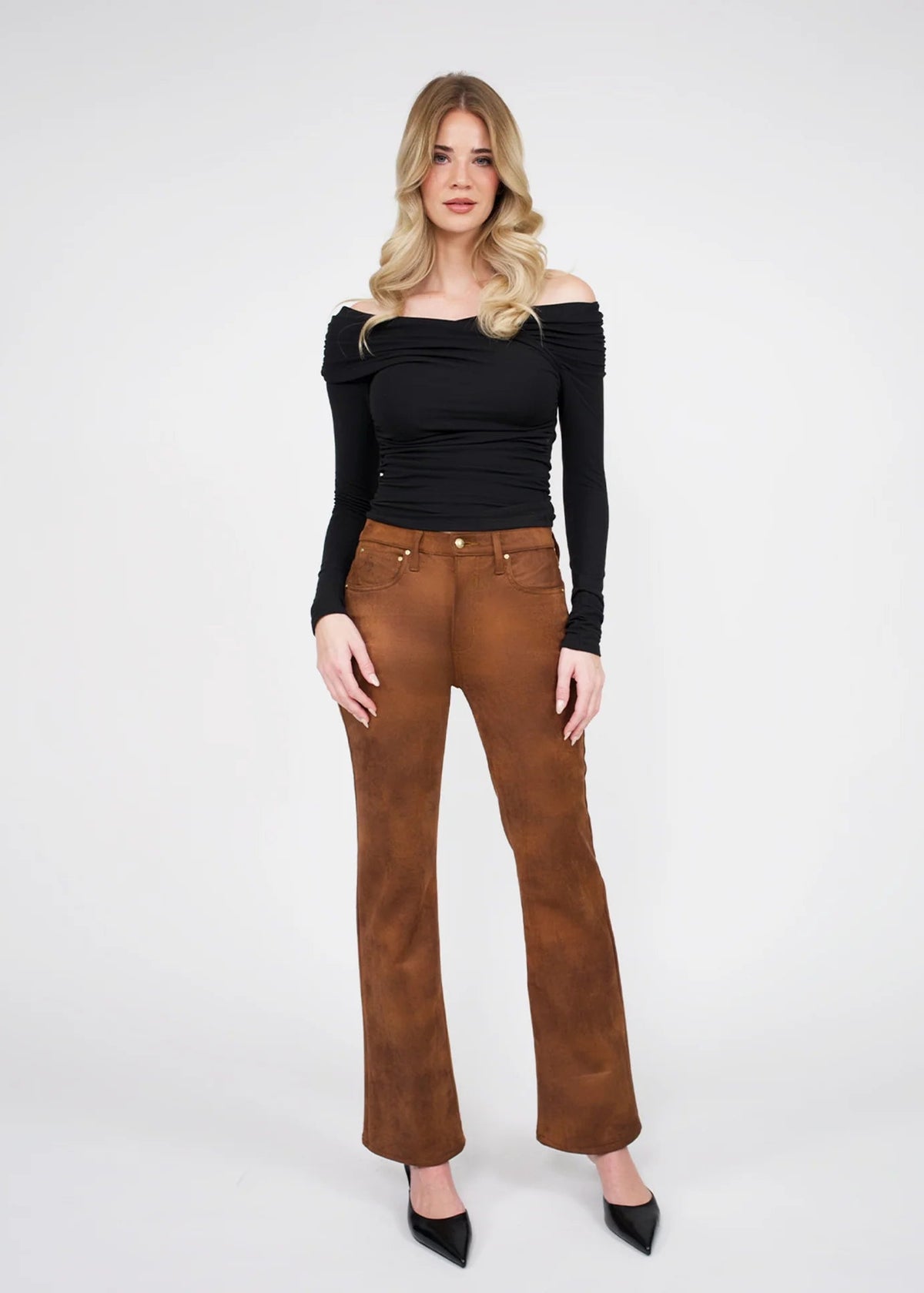 A woman modeling a black off-the-shoulder long-sleeve top and brown suede-like flare pants with black pointed heels, standing against a light gray studio background.