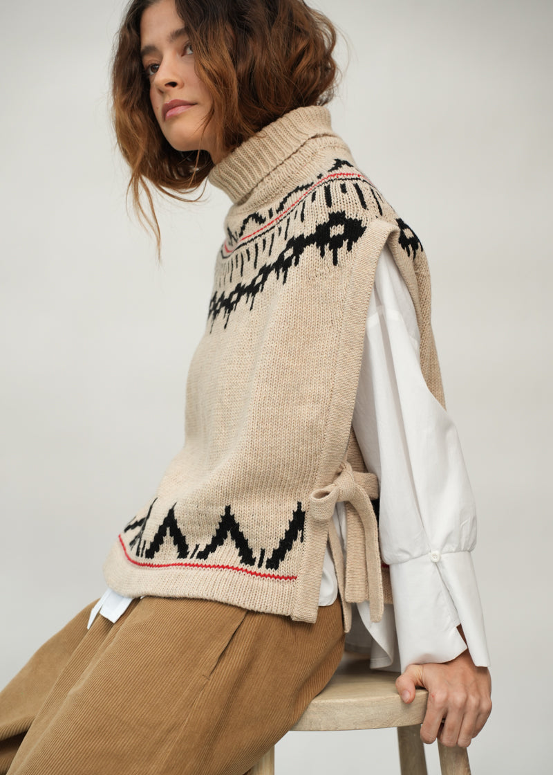 A woman seated on a wooden stool wearing a beige knit sweater vest with black geometric patterning and a thin red stripe, layered over a white long-sleeve top and tan trousers.