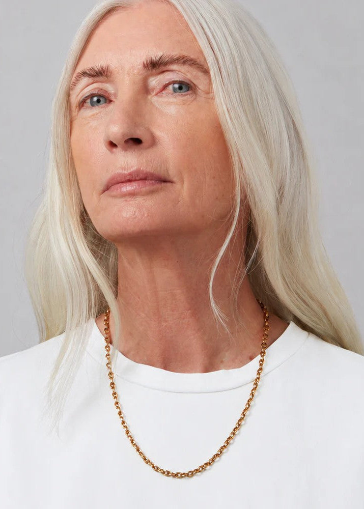 A close-up of a woman with platinum blonde hair wearing a delicate gold chain necklace and a white crewneck top, set against a neutral gray background.