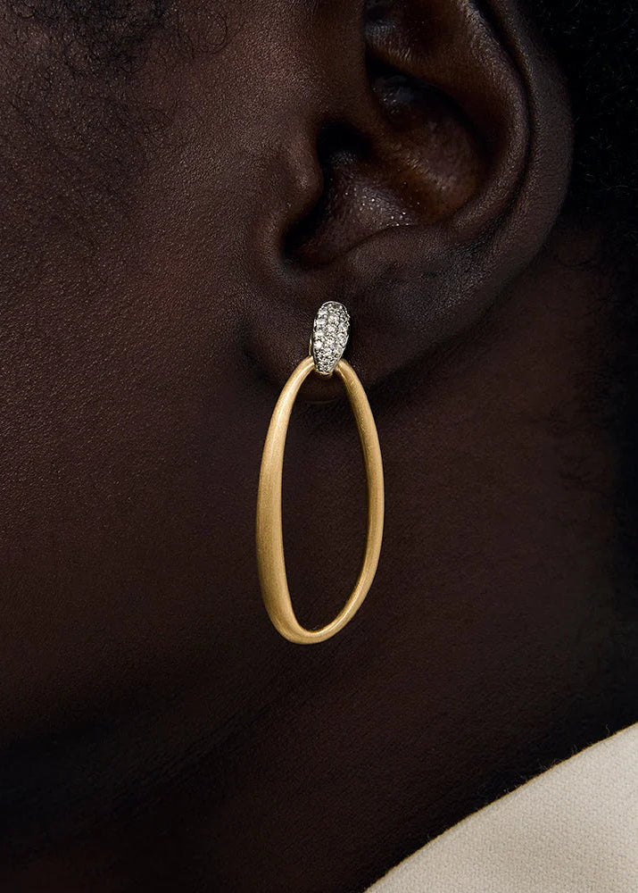 A close-up of a person's ear wearing a large gold hoop earring with a small pavé-set oval top, against a dark skin tone and light background.