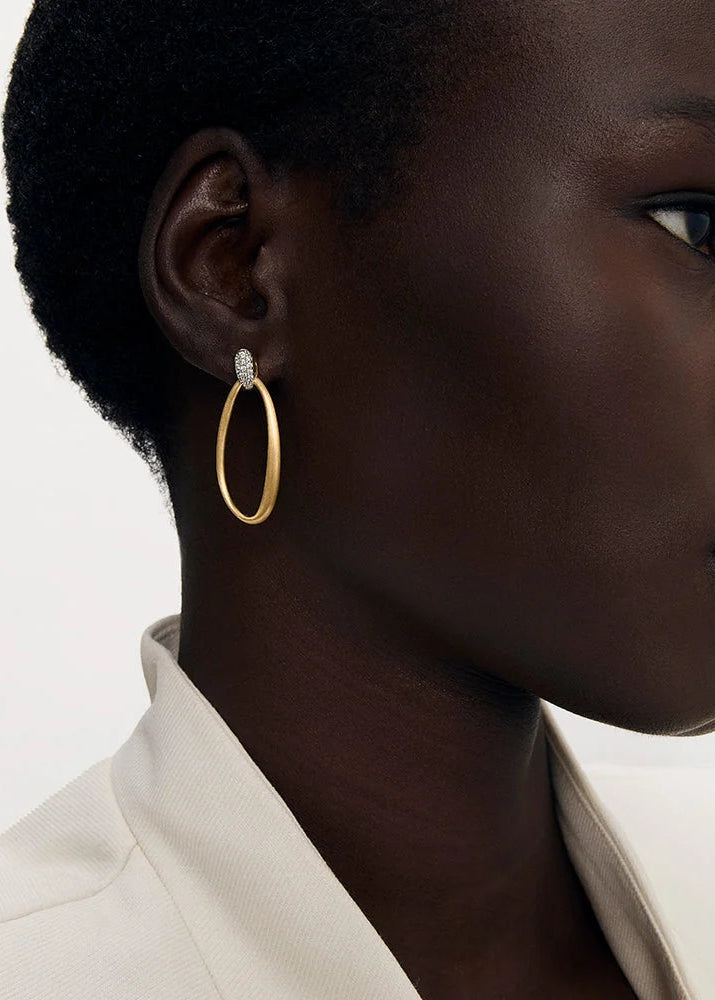 A close-up side profile of a model wearing a large, polished gold hoop earring with a small clear stone accent at the top, paired with a white collared shirt against a clean white background.