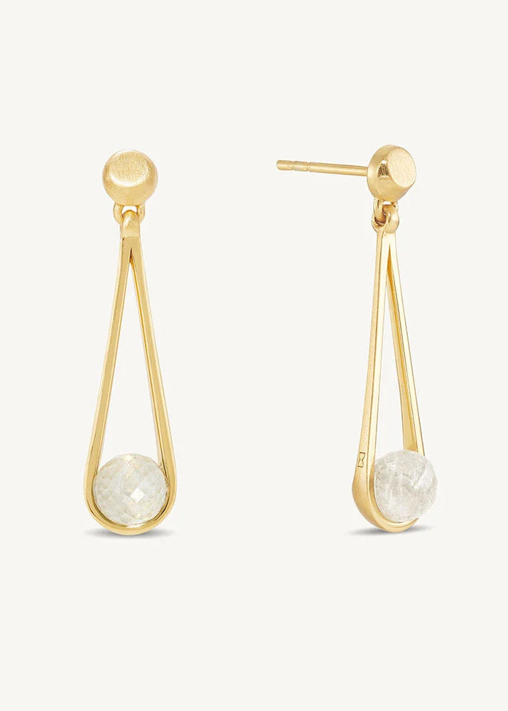 A pair of gold drop earrings with a circular faceted stone at the bottom, shown on a light cream background.