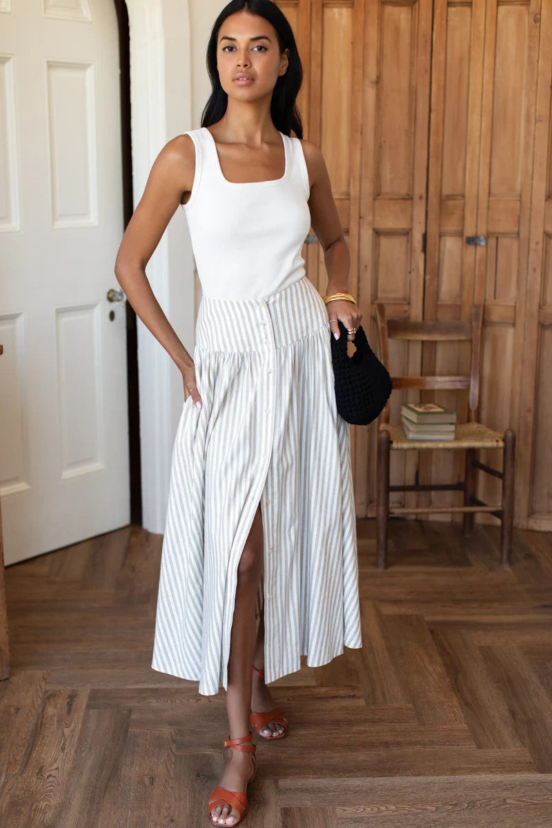 Woman in a white sleeveless top and striped skirt standing in a room with wooden flooring.