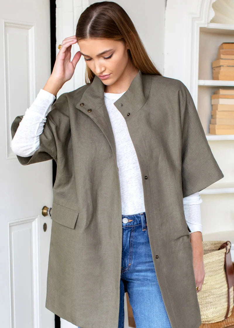 Woman wearing a long olive green coat over a white shirt and blue jeans, standing indoors.