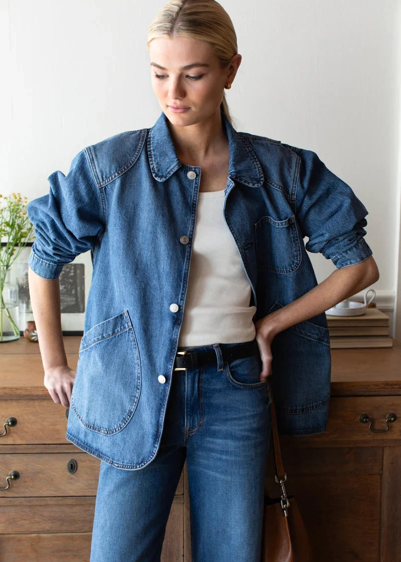 Woman wearing a denim jacket and jeans standing in a room with wooden furniture.
