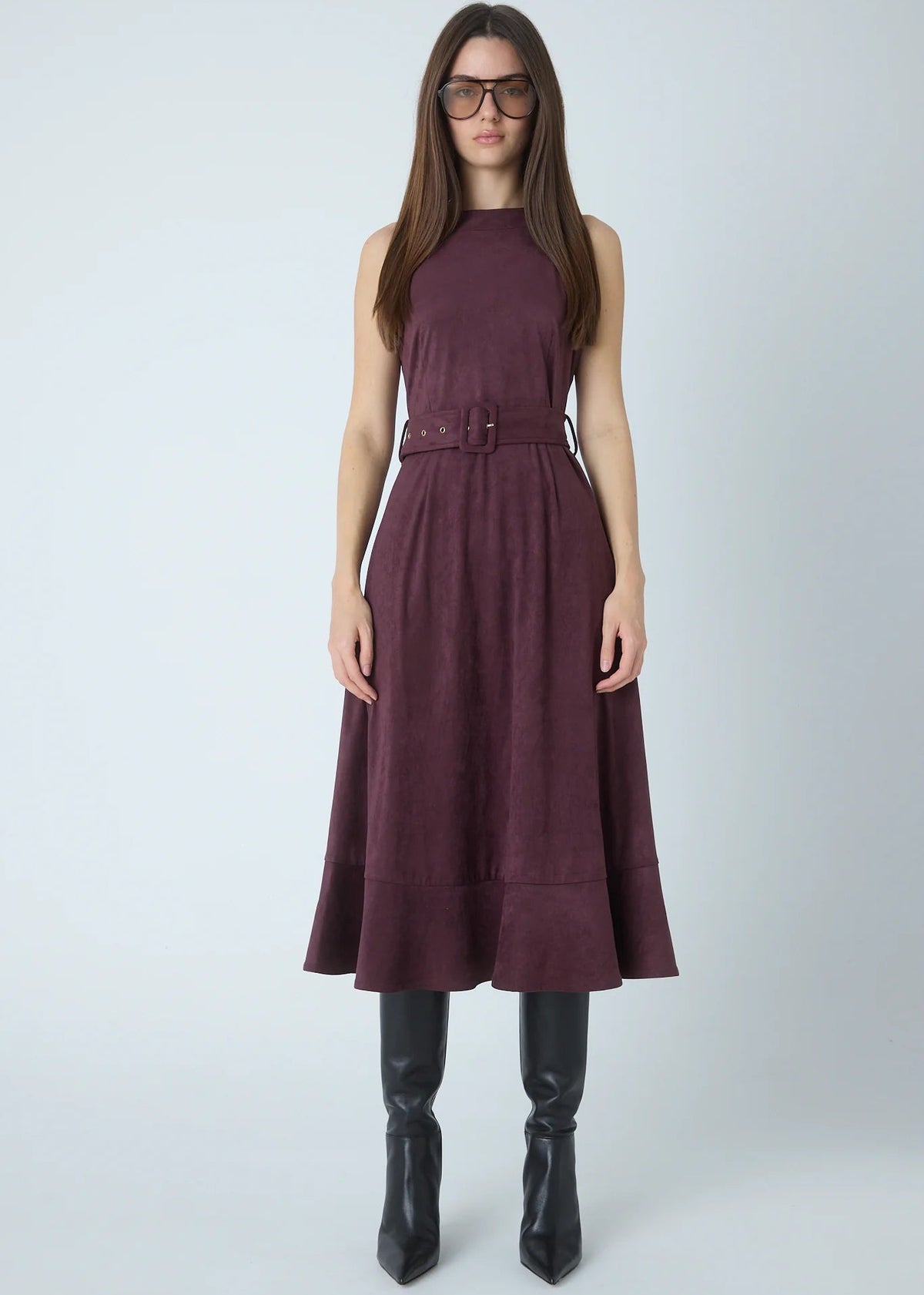 A woman modeling a sleeveless high-neck midi dress in a deep plum purple color, featuring a matching belt at the waist. She is also wearing black knee-high leather boots and dark sunglasses, standing against a light gray studio background.