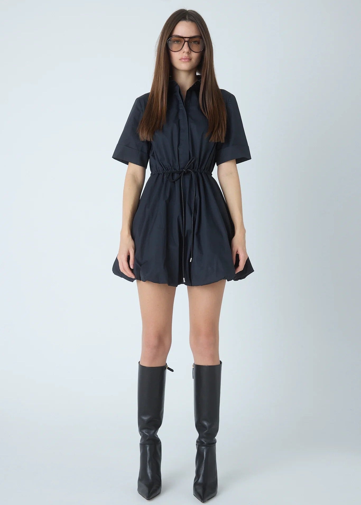 A woman modeling a dark navy short-sleeve mini dress with a drawstring waist, paired with black knee-high boots, against a light gray studio background.