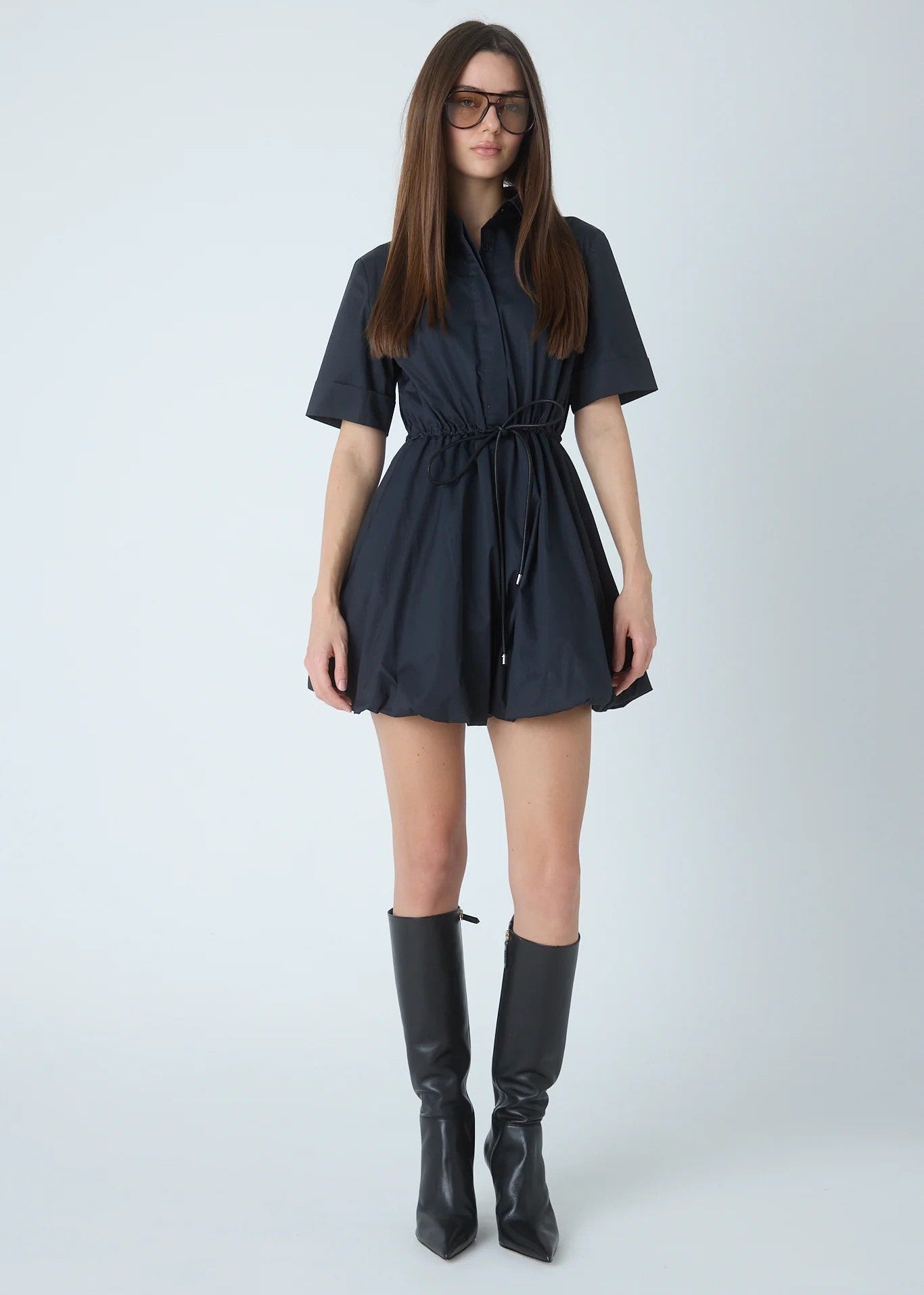 A woman modeling a dark navy short-sleeve mini dress with a tie waist, paired with black knee-high boots, against a light gray studio background.