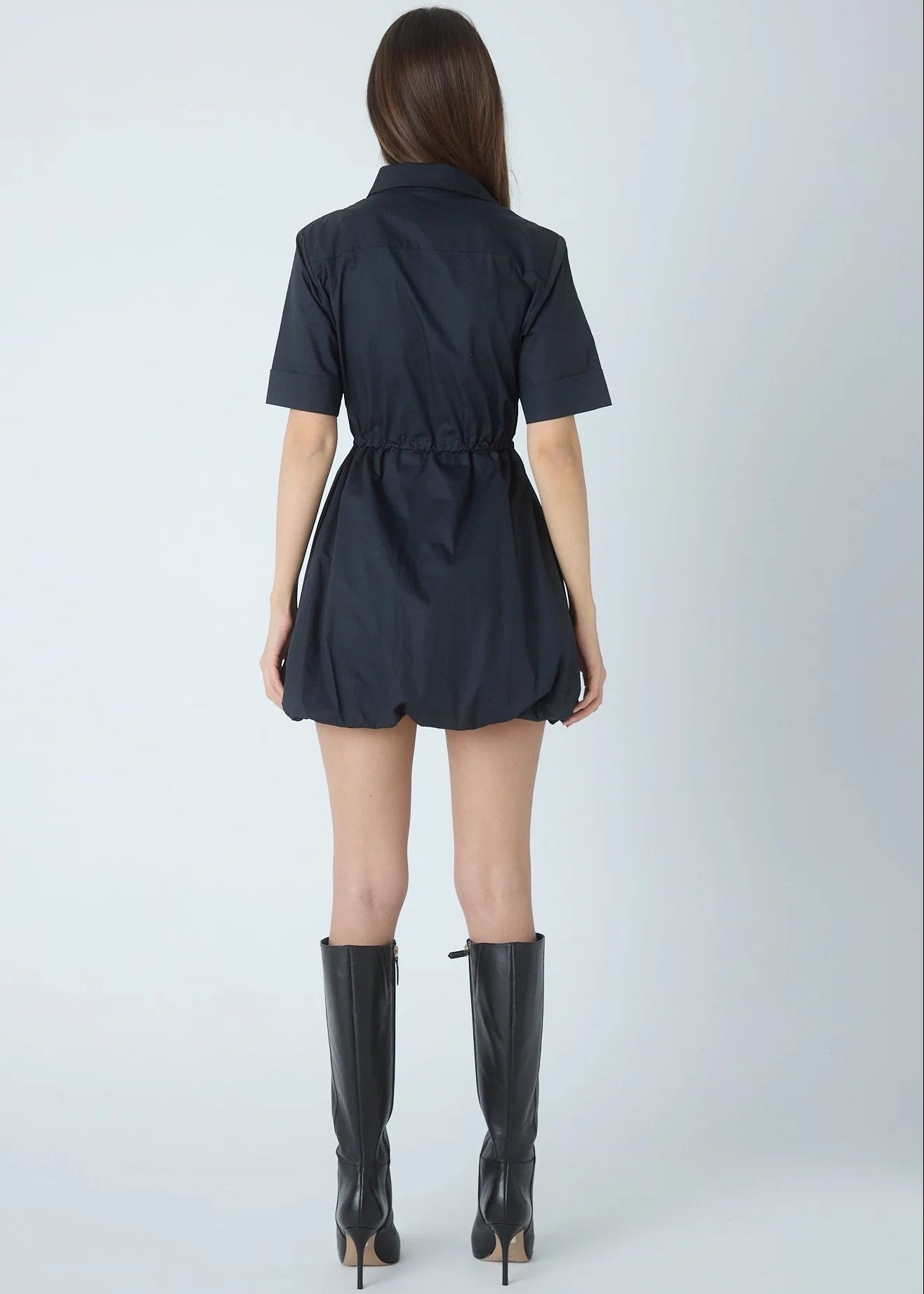 A woman shown from the back wearing a short-sleeve dark navy mini dress with a cinched waist and black knee-high heeled boots, photographed against a light gray studio background.