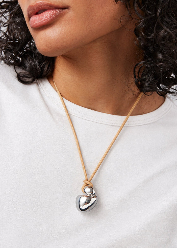 A close-up of a person wearing a silver heart pendant necklace on a tan cord, paired with a white t-shirt.