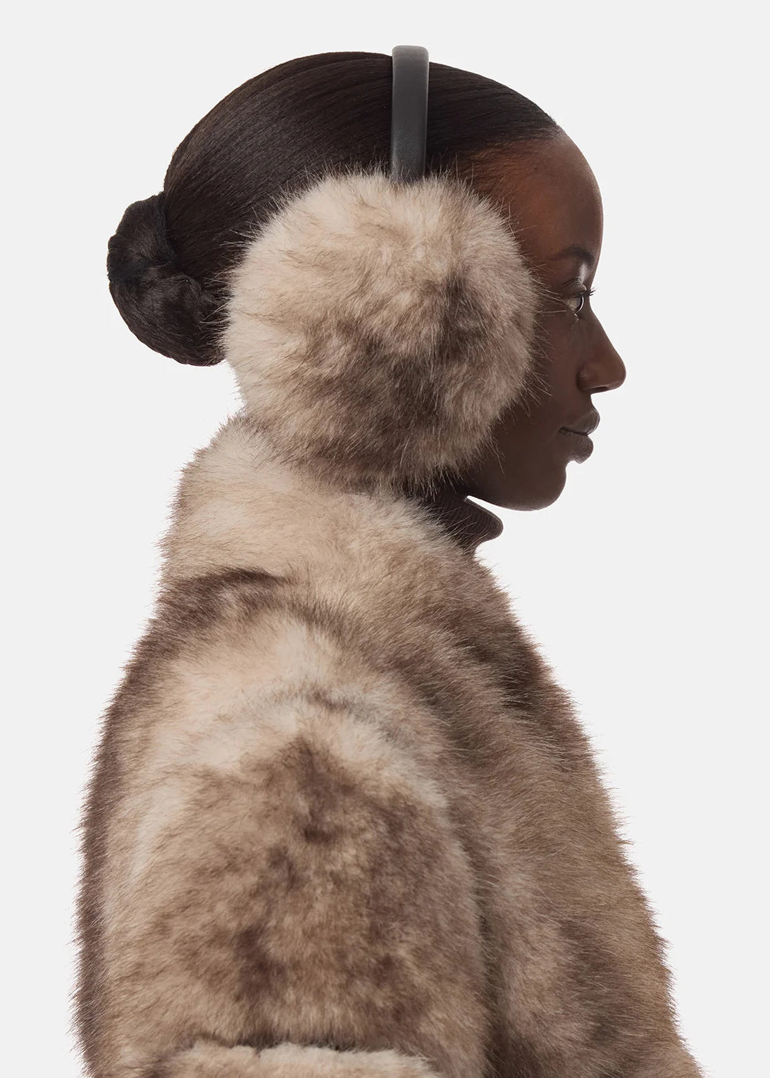 A side-profile portrait of a model wearing plush faux-fur earmuffs with a dark headband and a matching beige-brown fur coat, set against a solid light gray background.