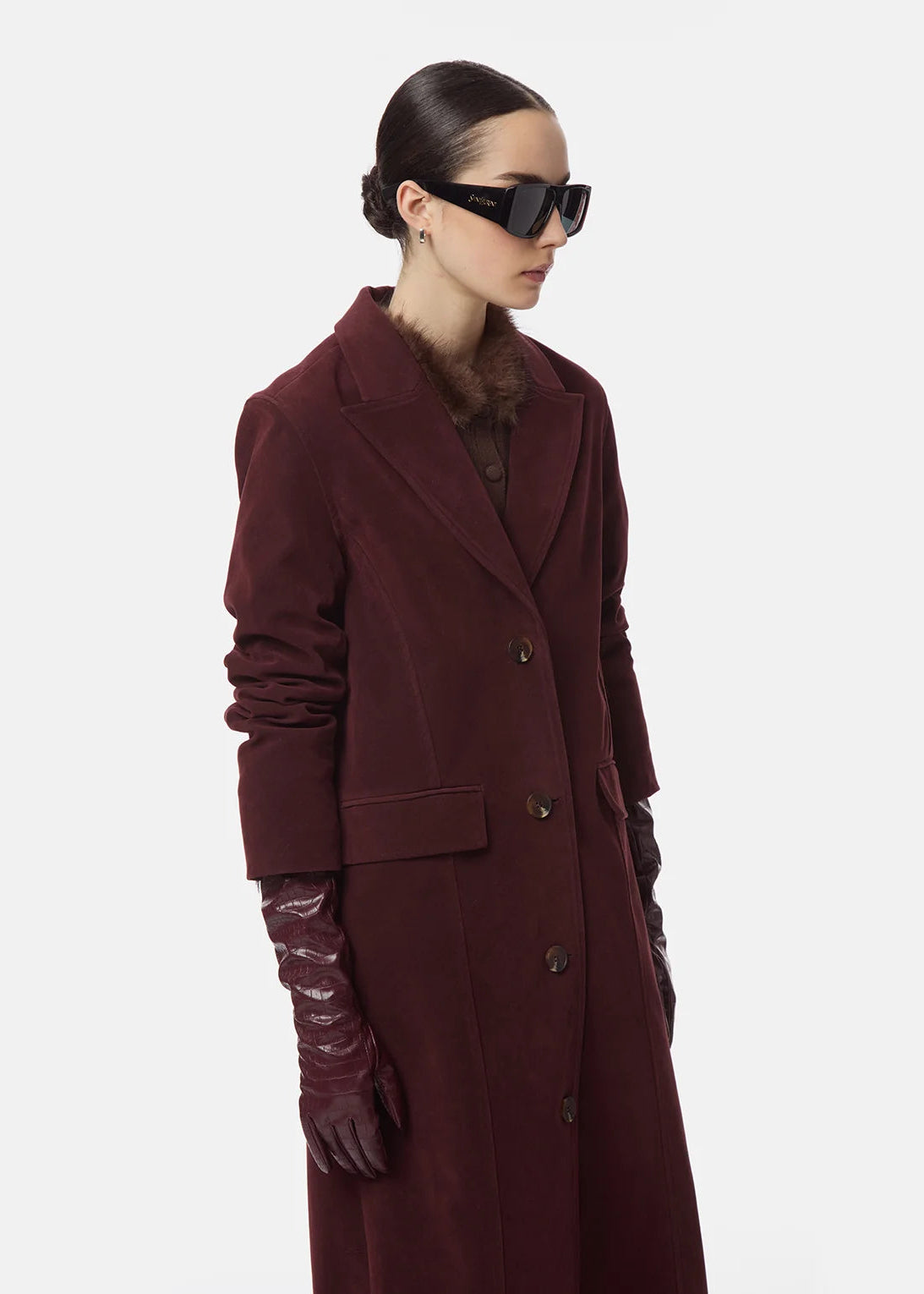 A model wearing a long burgundy wool-blend coat with structured shoulders, dark brown leather gloves, and black sunglasses, shown from a side profile against a light gray studio background.