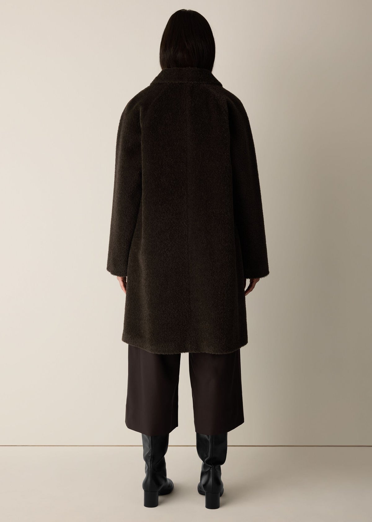 A model viewed from the back wearing a dark brown textured coat, wide-leg dark brown trousers, and black ankle boots against a plain beige studio background.