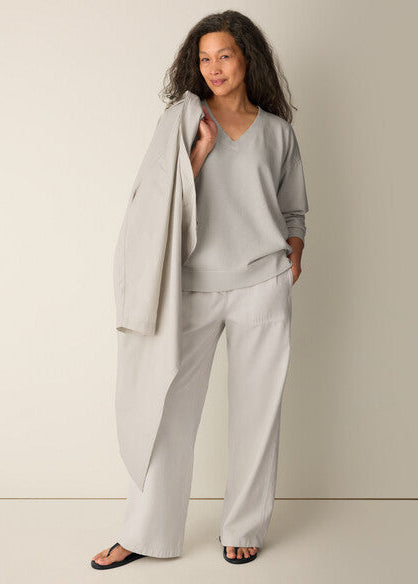 A woman modeling a light gray lounge set consisting of a V-neck long-sleeve top and matching wide-leg pants, with a draped light gray cardigan over her shoulder, set against a plain beige studio background.