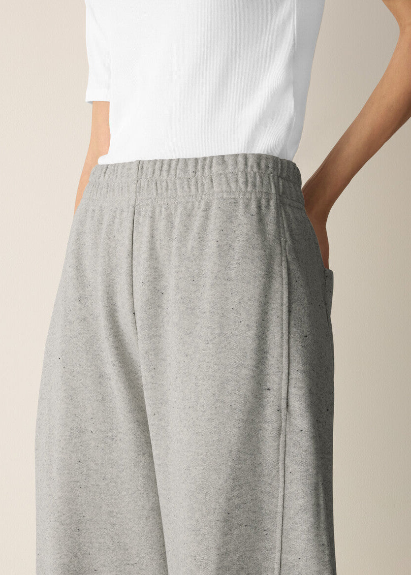 Close-up of a person wearing light heather gray wide-leg sweatpants with an elastic waistband and side pockets, paired with a white top against a plain beige studio background.