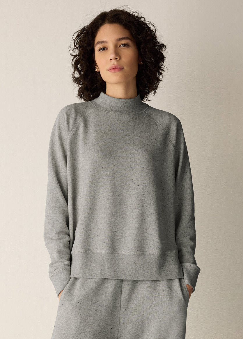 A woman with curly dark hair wearing a light gray mock neck sweatshirt and matching gray sweatpants, standing against a plain light beige studio background.
