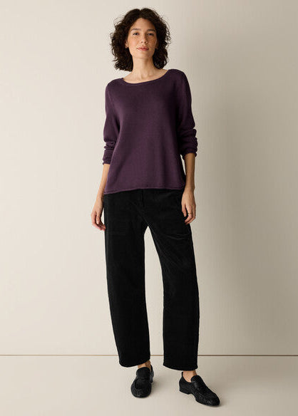 A woman modeling a dark purple long-sleeve knit top and black wide-leg trousers against a plain beige studio background.